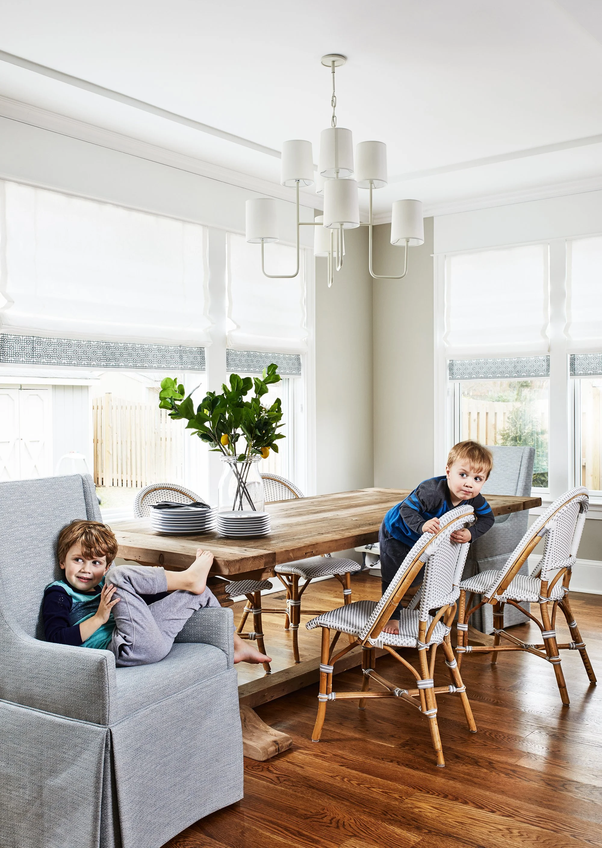 Bright dining room with a wood table, woven chairs, gray upholstered host chair, white chandelier, leafy branches, and Roman shades.