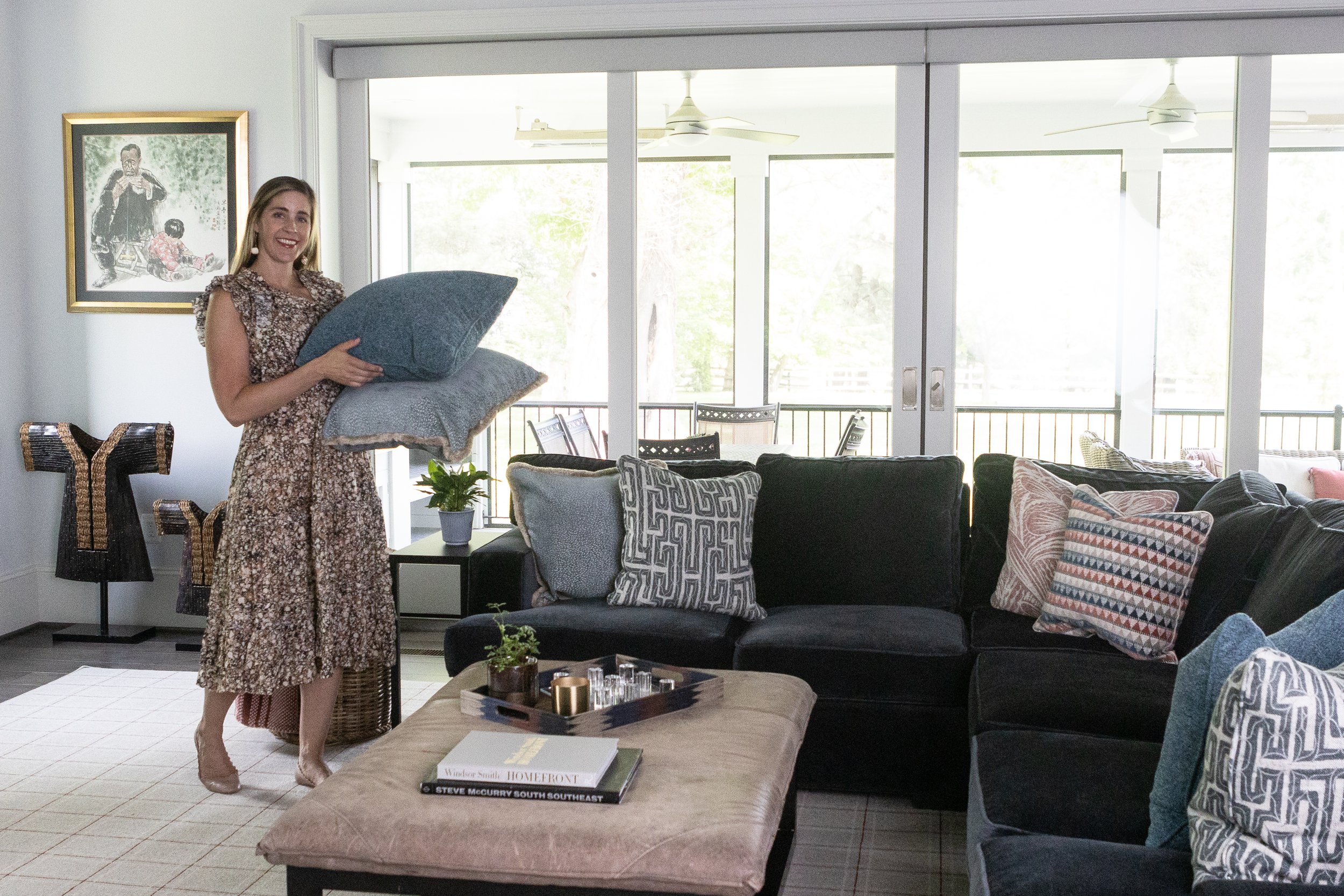 Regan styling patterned pillows on a dark sectional in a living room with large windows, framed artwork, plants, and books.