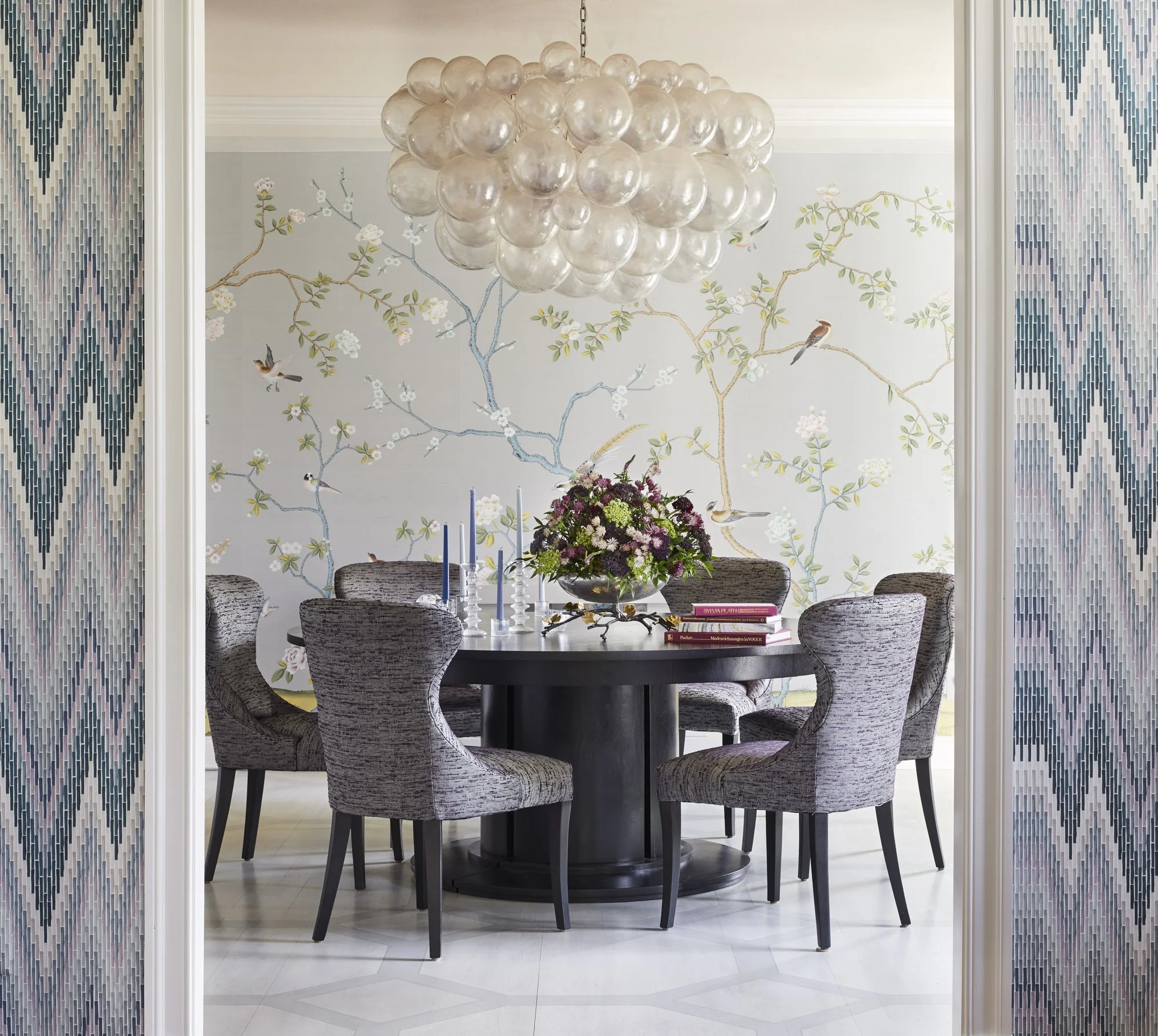 Dining room viewed through a wide doorway framed by textured wallpaper, with a round table, upholstered chairs, clustered pendant light, and floral bird wallpaper.