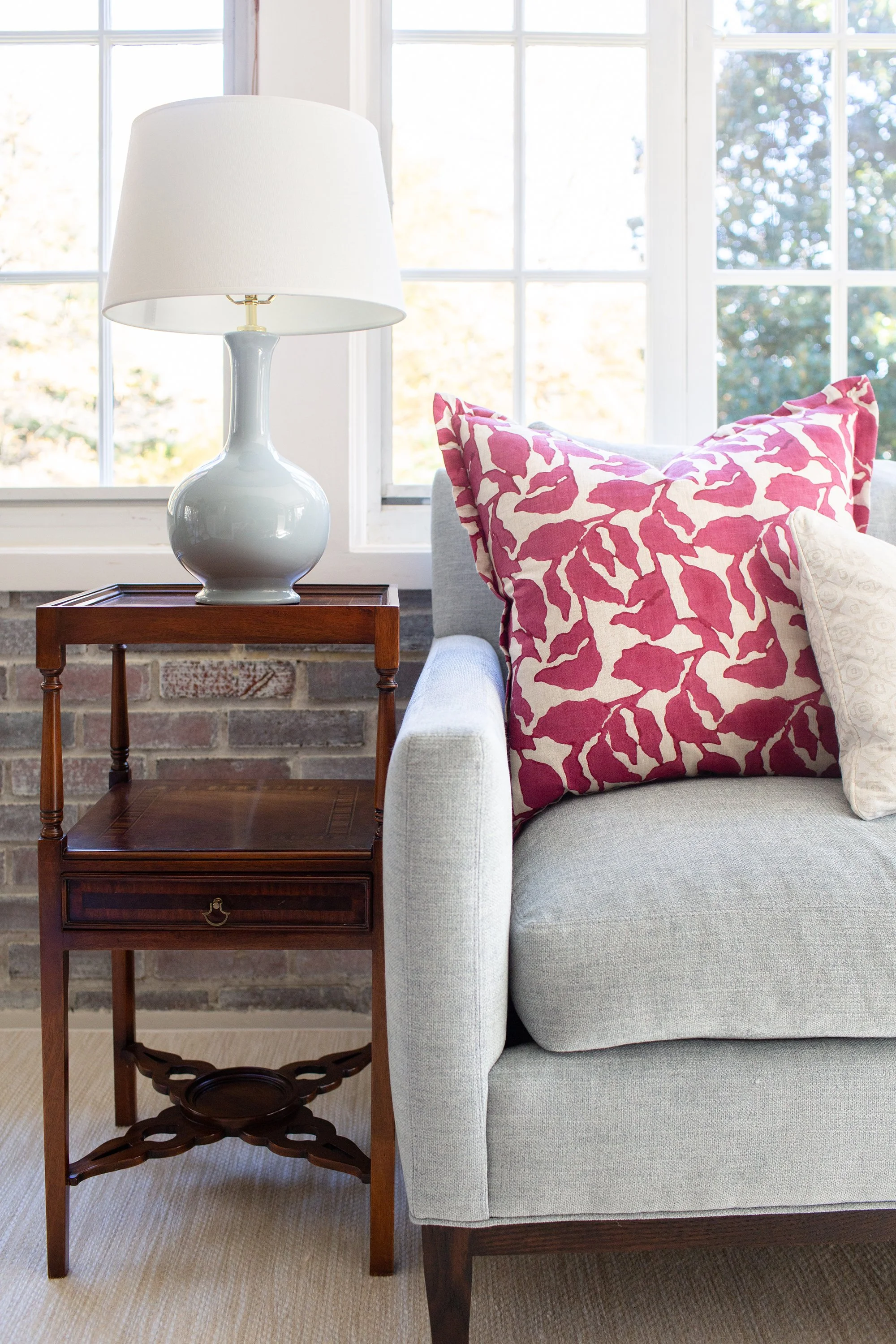 Light gray sofa with a pink patterned pillow beside a wood table and pale blue lamp near a large window.