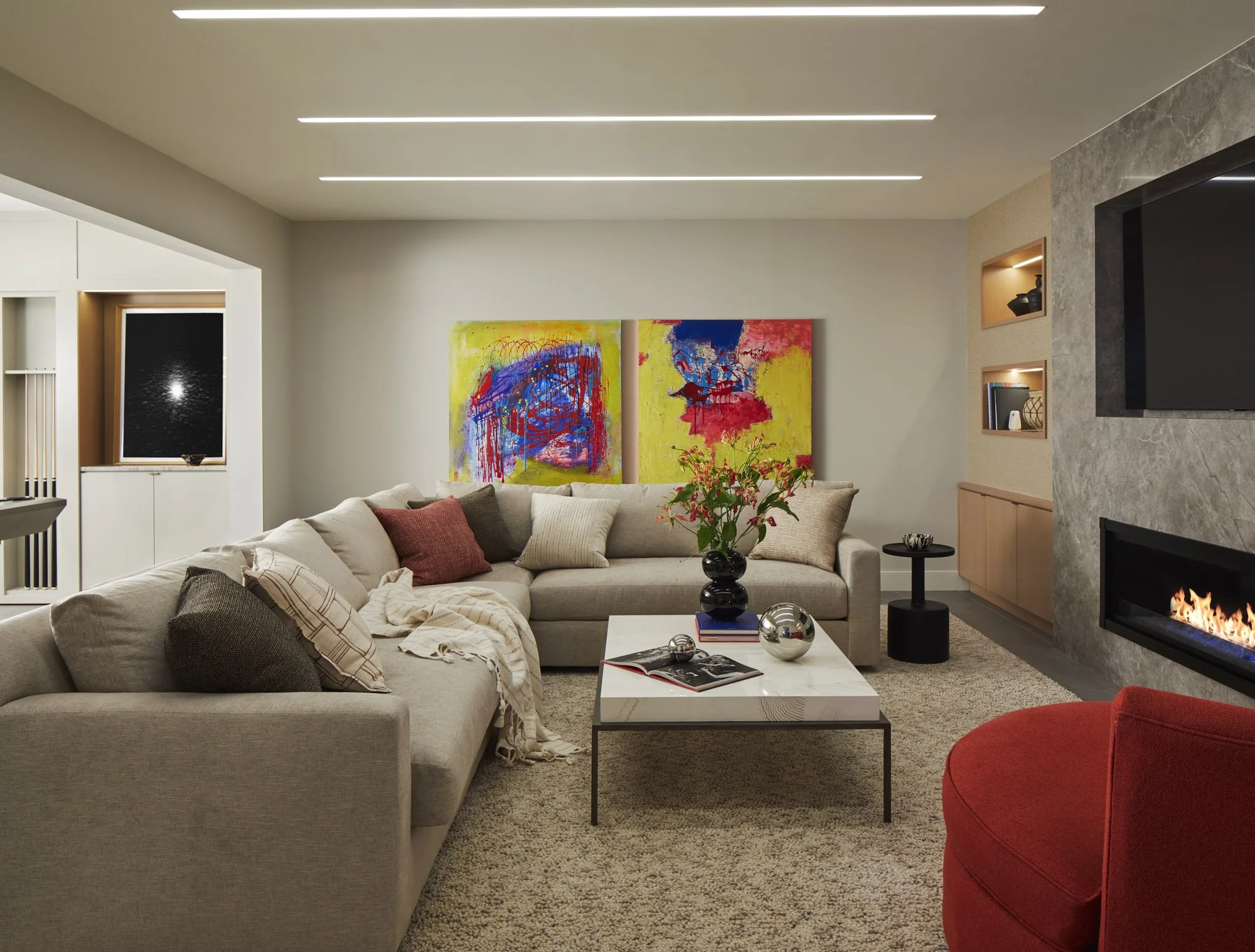 Cozy movie lounge with a gray sectional, colorful abstract artwork, a marble coffee table, red chair, and fireplace.
