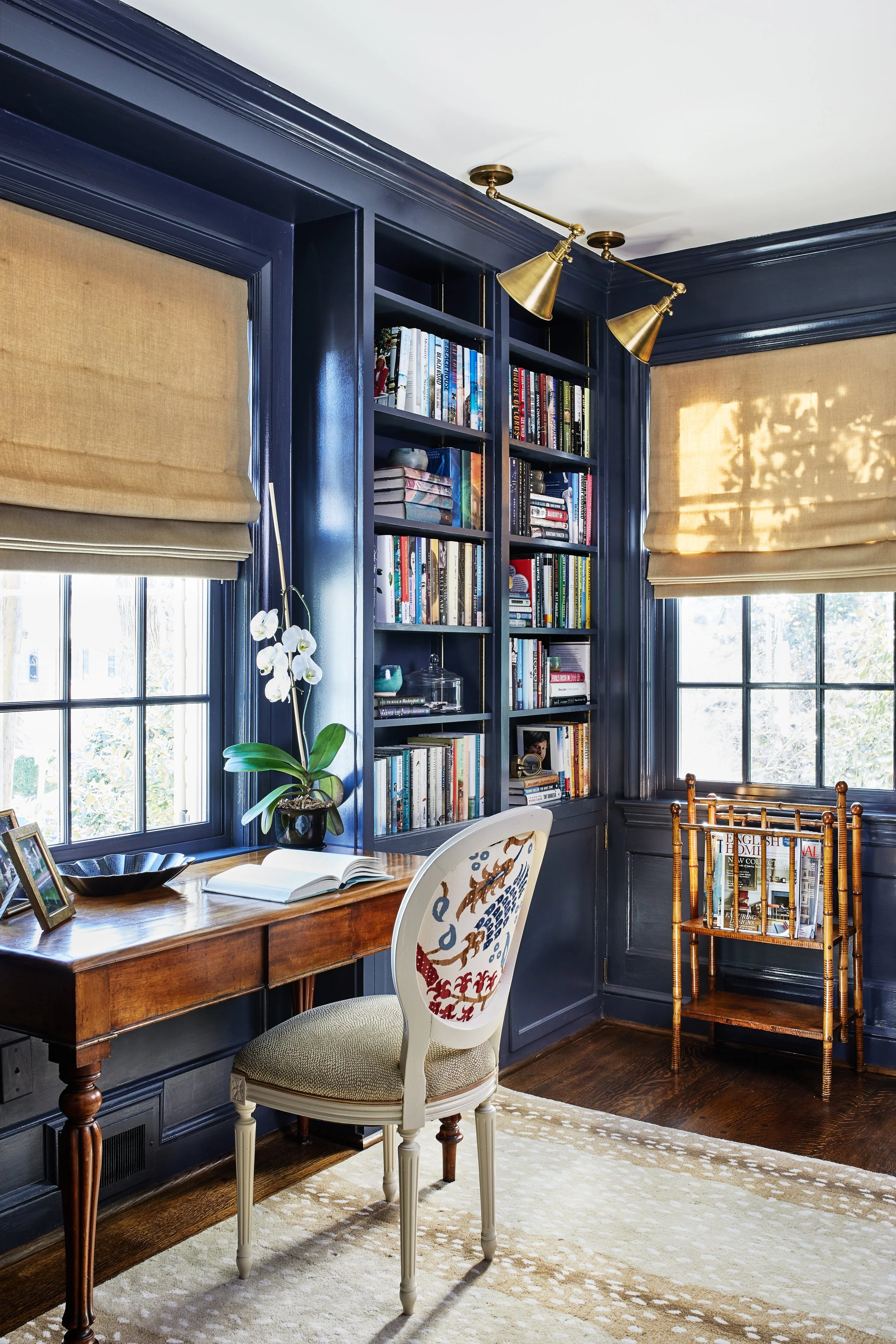 Navy home office with built-in bookshelves, brass lighting, woven shades, an antique desk, and an upholstered chair.