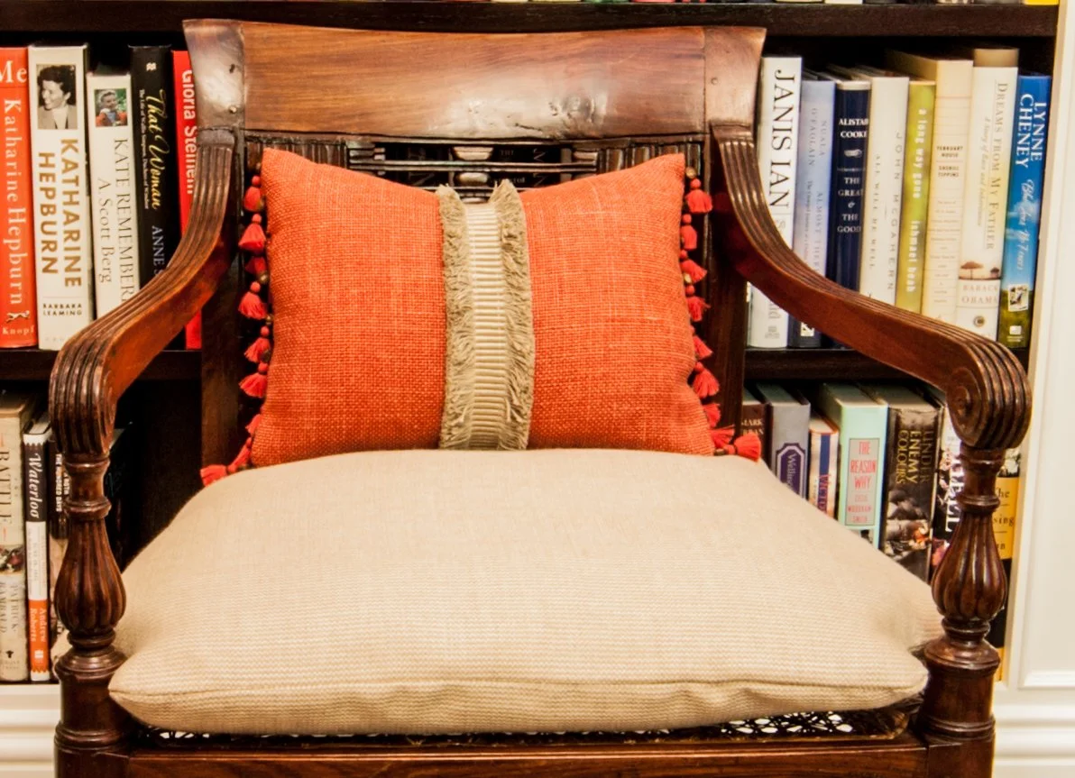 Wood armchair with red patterned pillows positioned in front of a book-filled shelf.