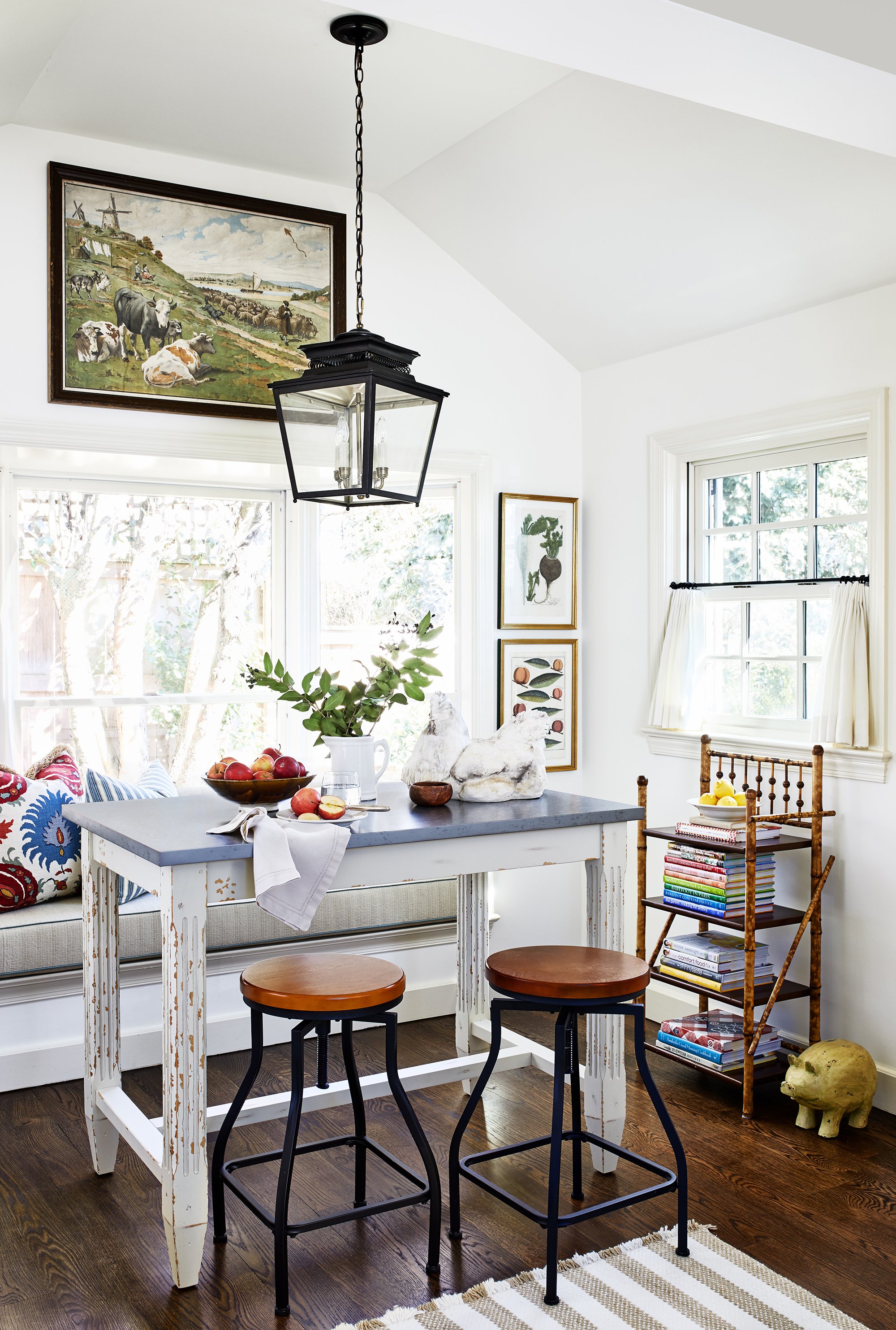 Cozy kitchen nook with a small table, two stools, window seat, large painting, pendant light, books, and a decorative pig.