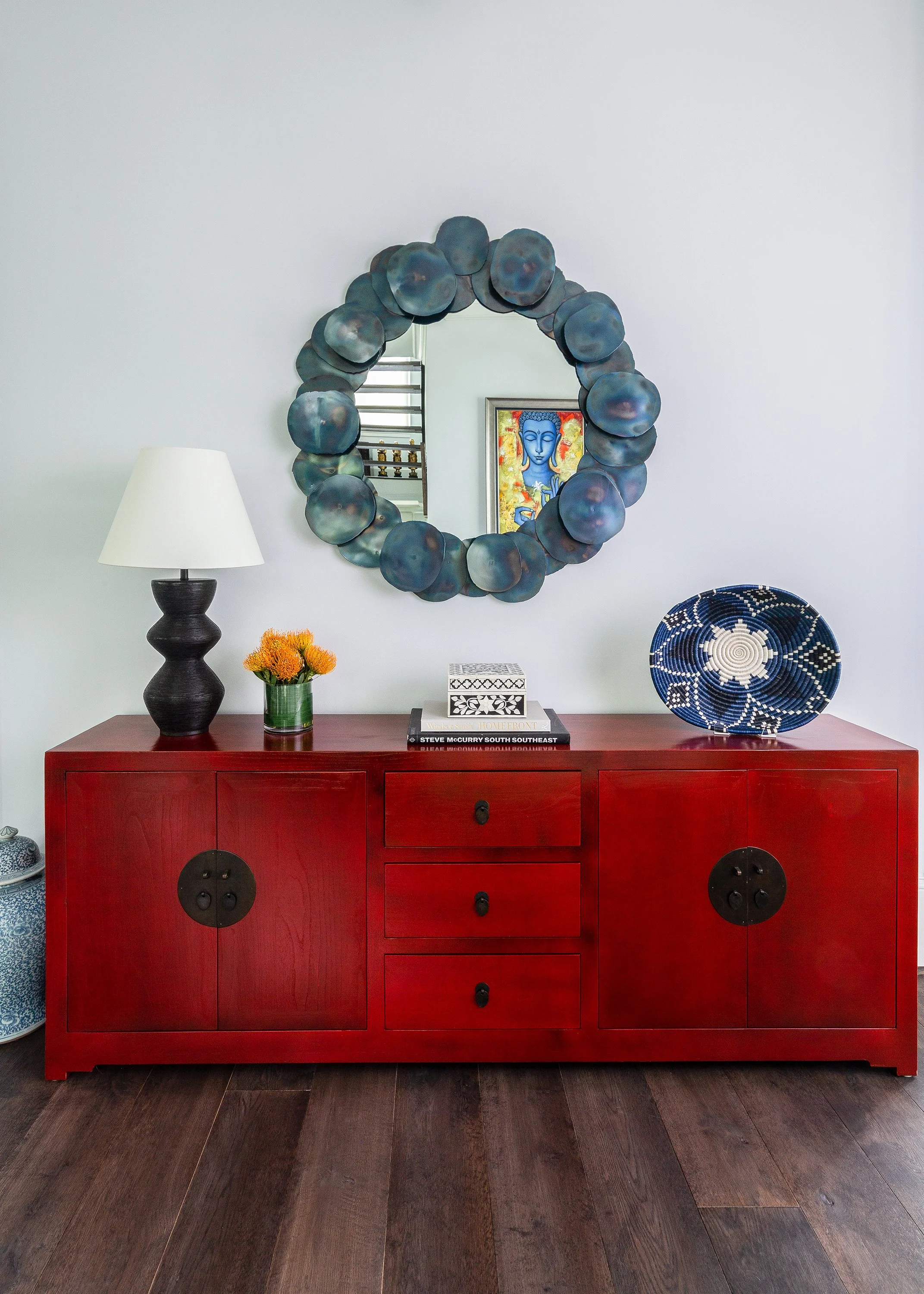 Red wood sideboard styled with a decorative mirror, blue-and-white bowl, table lamp, orange flowers, and books.