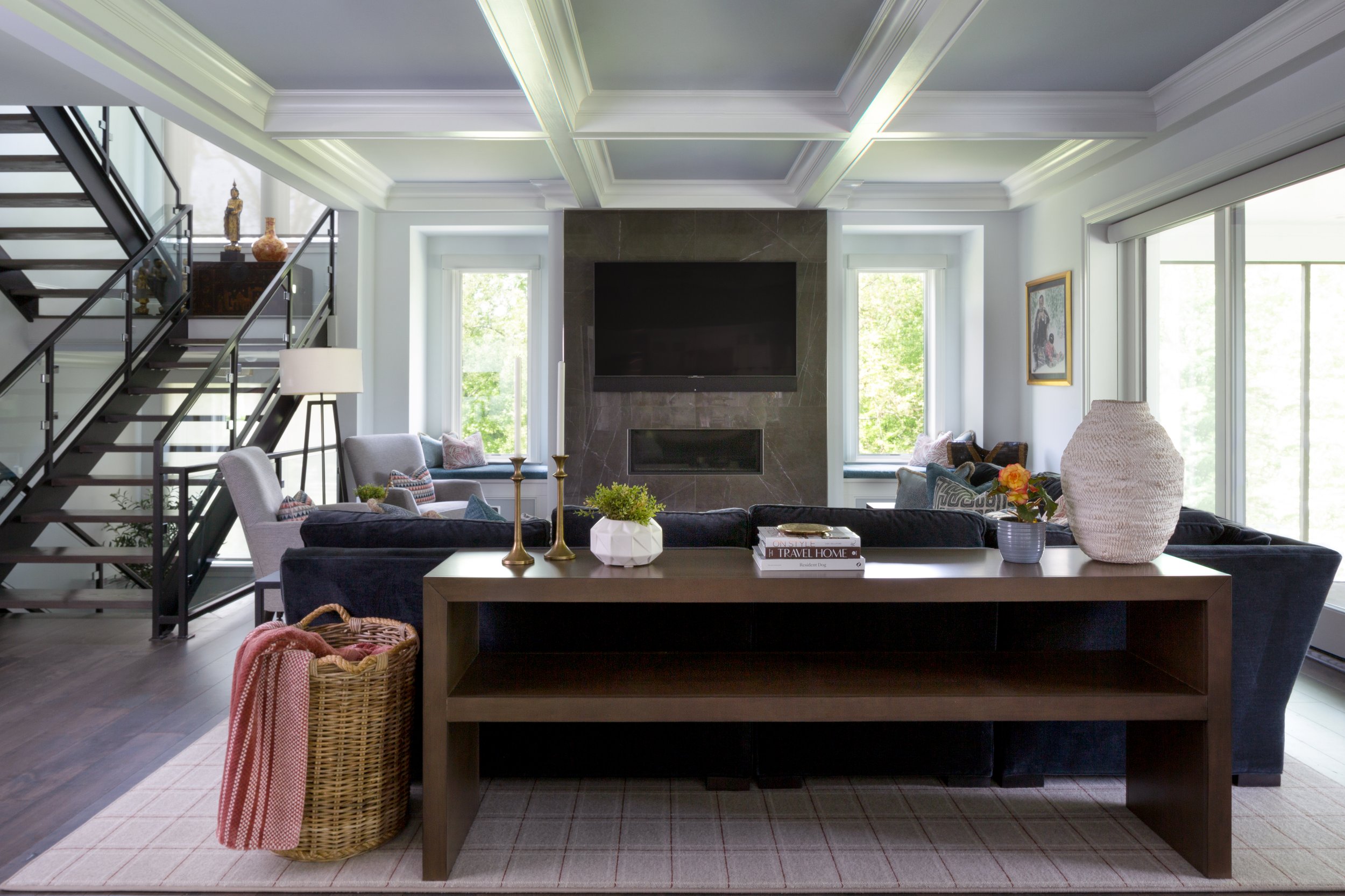 Spacious family room with a coffered ceiling, sofas, dark wood furniture, a stone television wall, black staircase, and large windows.