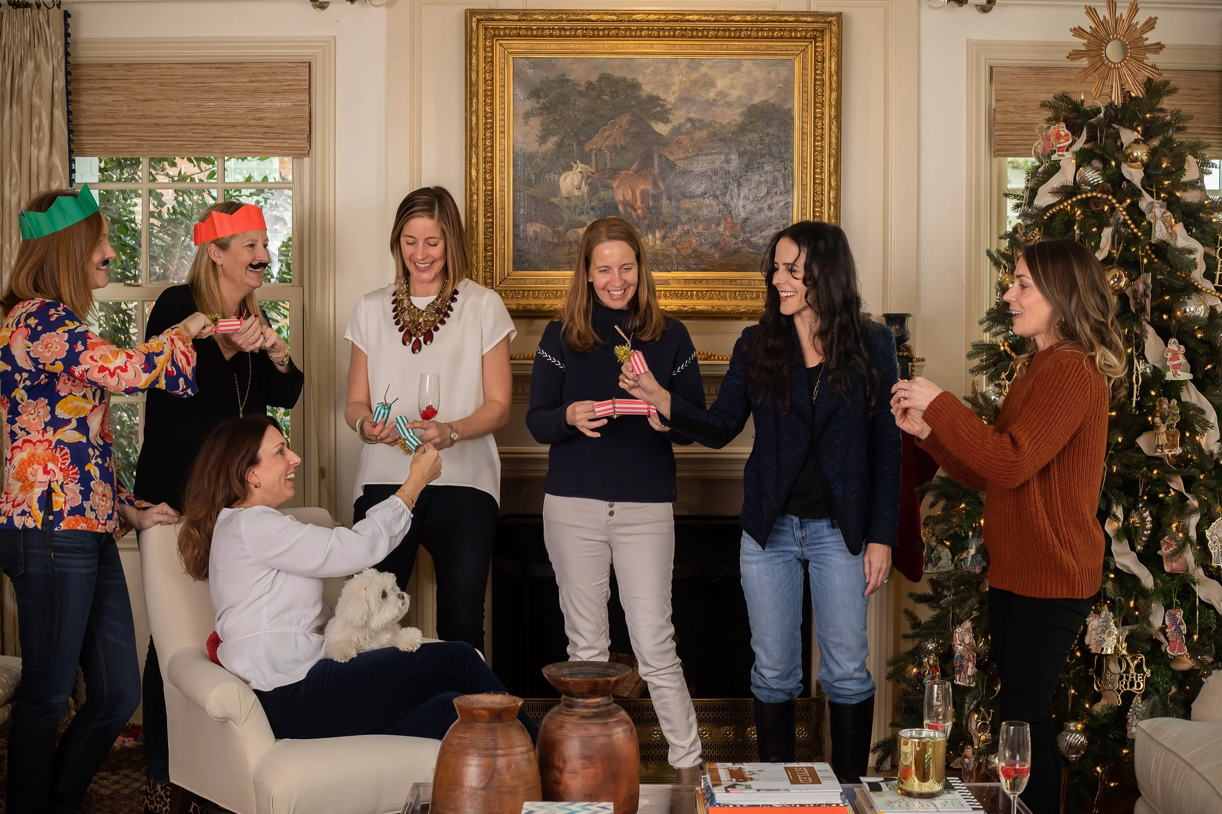 Women celebrating the holidays in a living room with a decorated Christmas tree, colorful artwork, and a small dog.