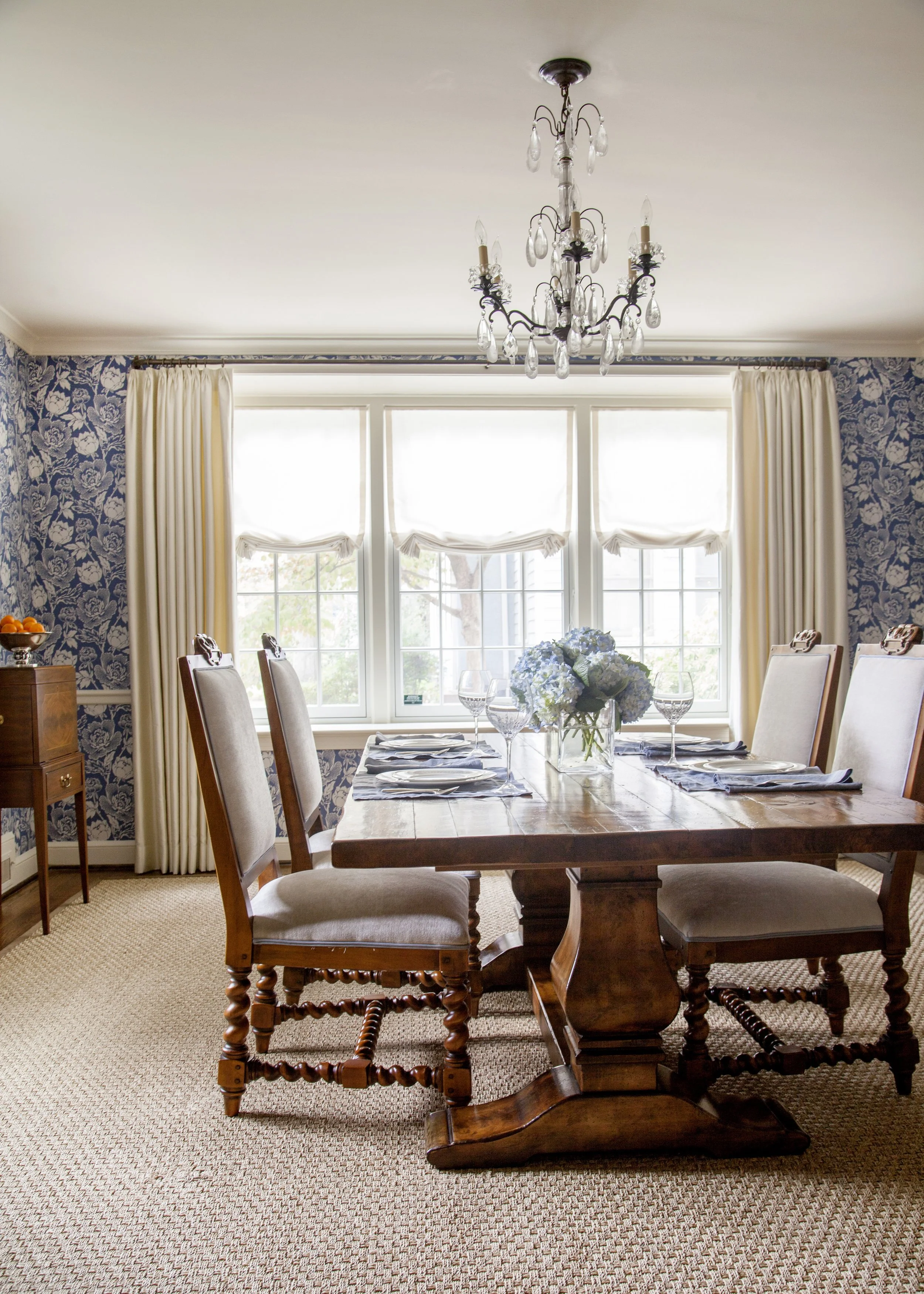 Elegant dining room with a wood table, upholstered chairs, chandelier, floral wallpaper, and blue flowers.