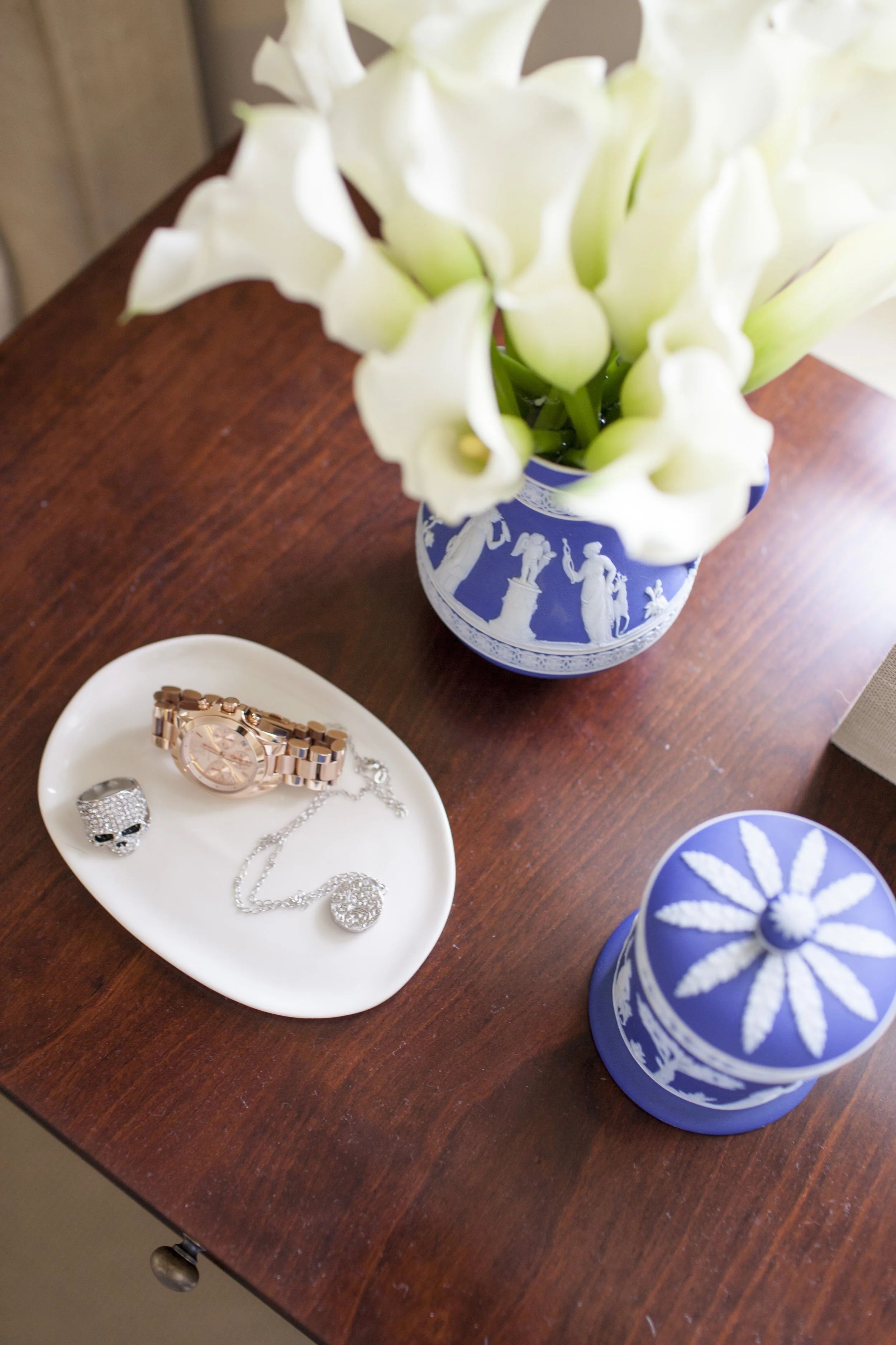 Wood table styled with white calla lilies in a blue vase and a tray holding a watch, ring, and necklace.
