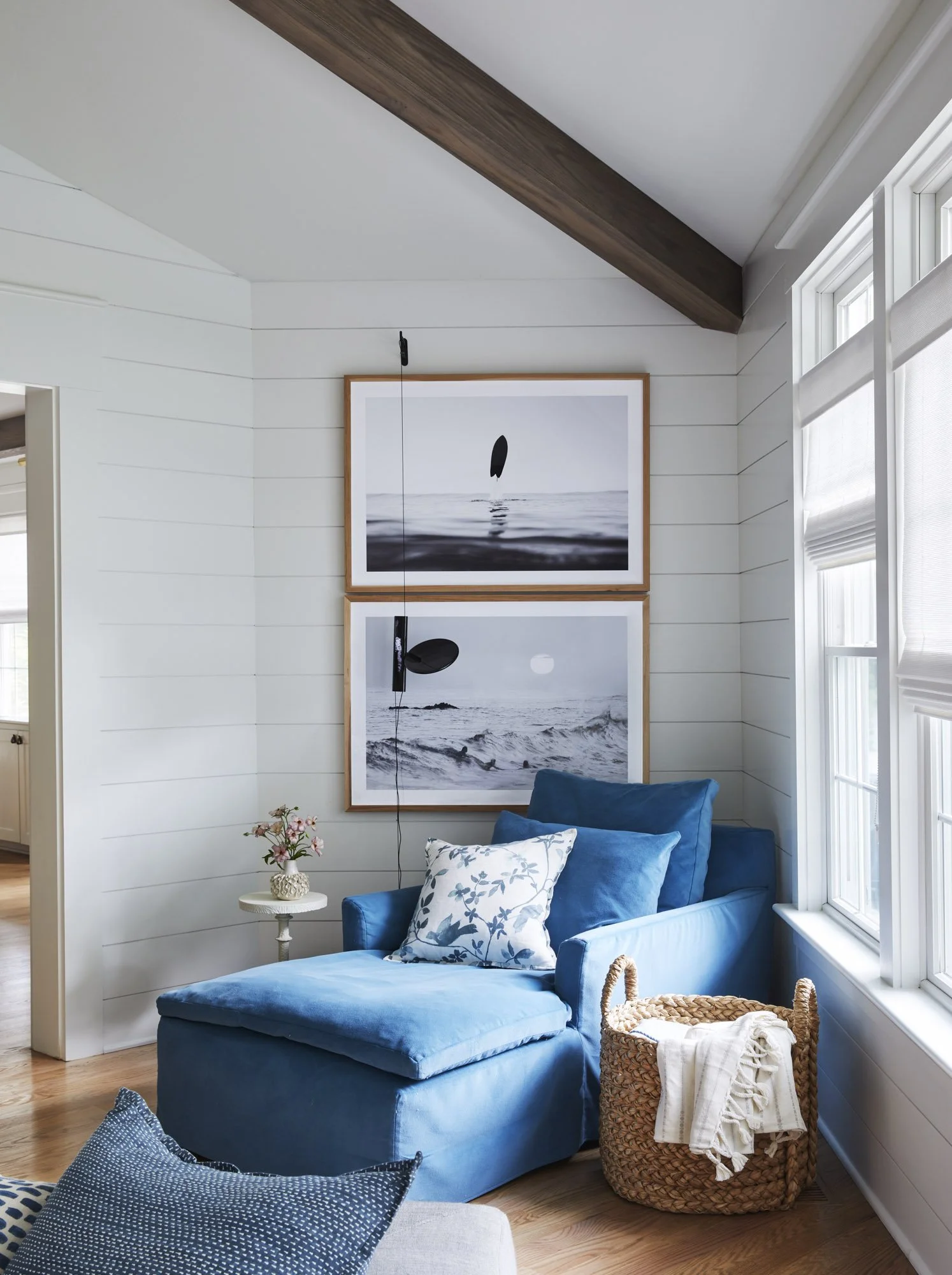 Coastal reading nook with a blue chaise, patterned pillows, framed ocean prints, shiplap walls, and a woven basket.