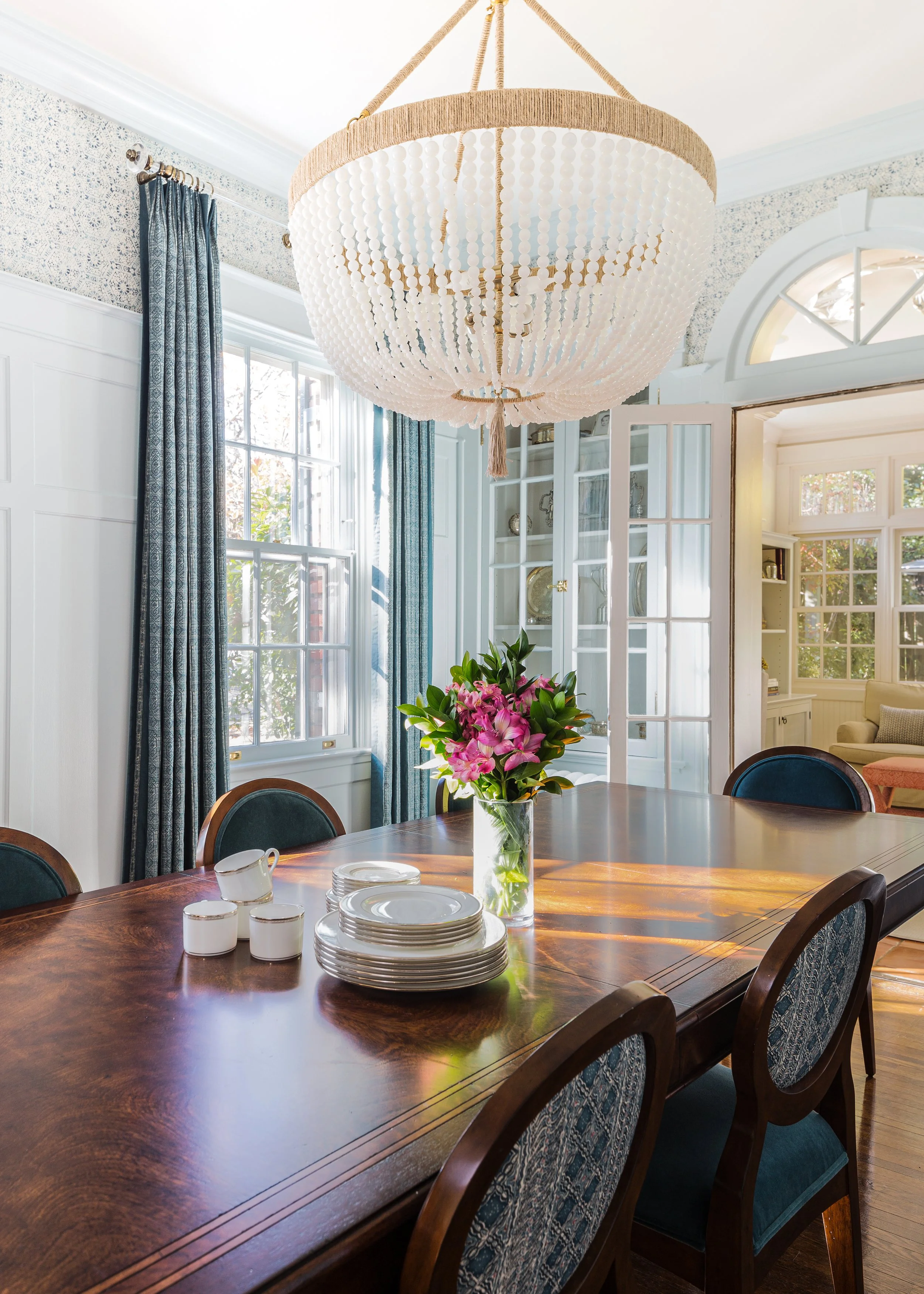 Formal dining room with a wood table, blue upholstered chairs, glass-front cabinets, chandelier, flowers, and candles.