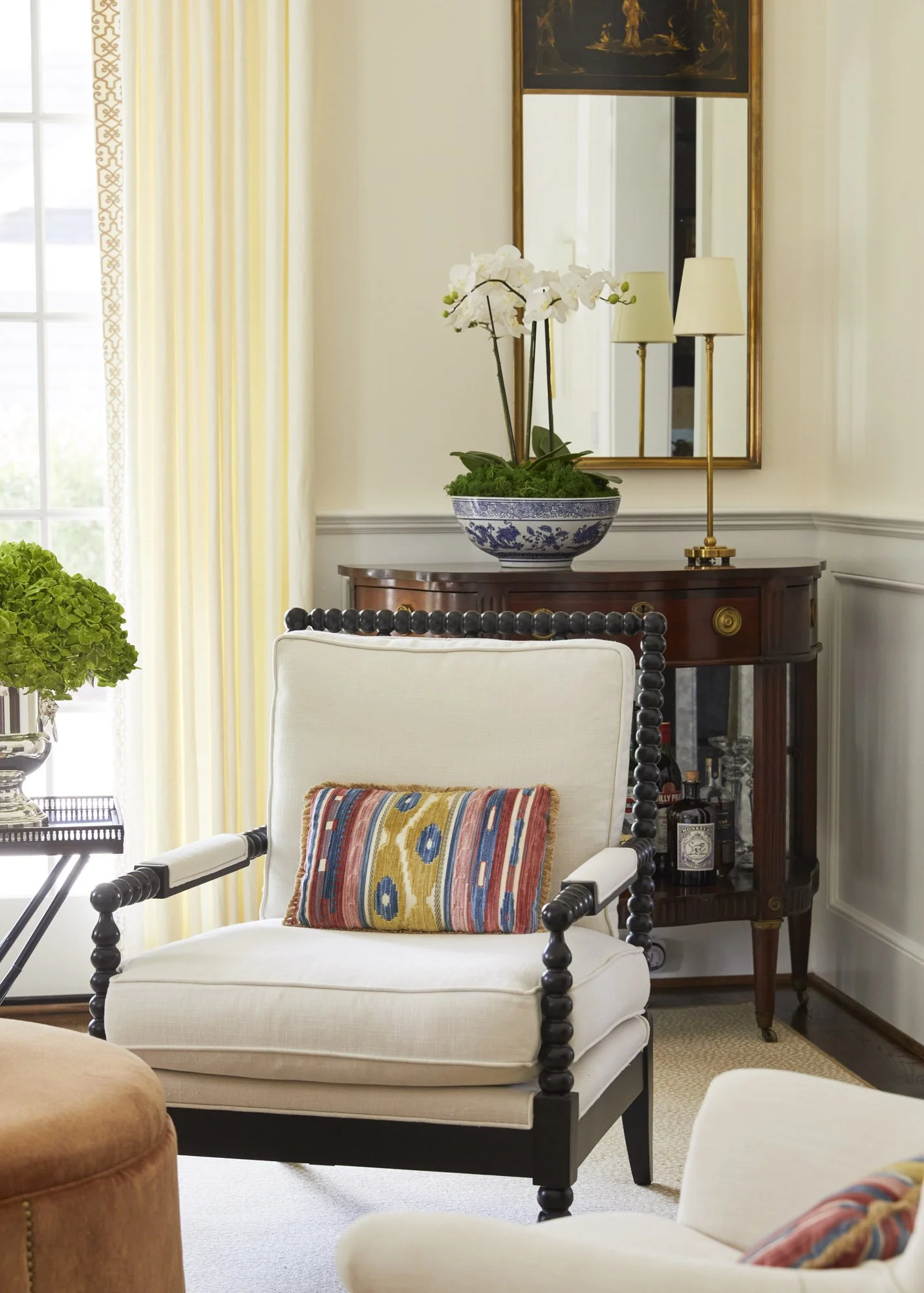 Black cat perched on a white armchair with a colorful pillow beside a potted plant, mirror, and tall floor lamp.