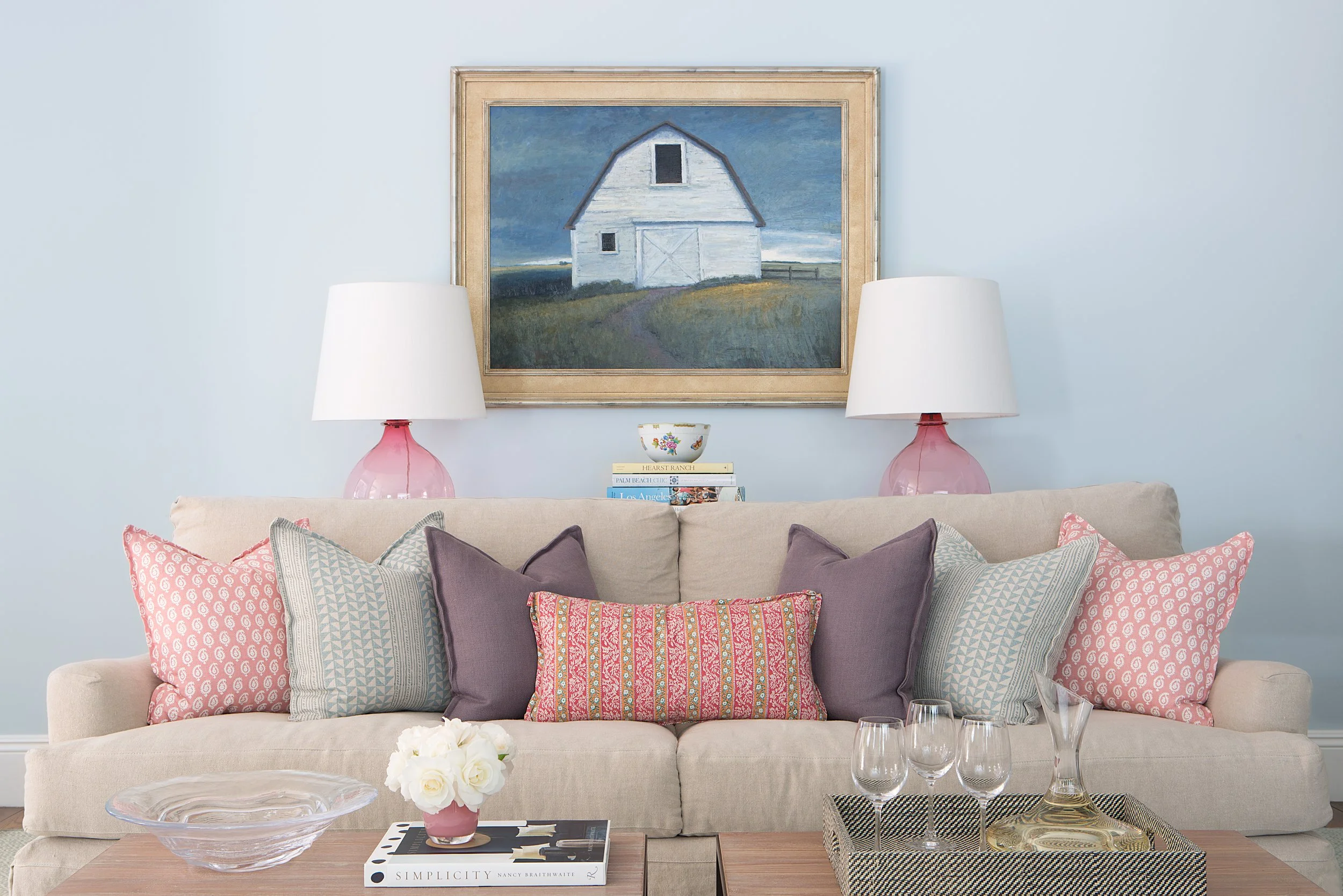 Beige sofa with colorful pillows beneath a barn painting, flanked by pink table lamps and a coffee table with flowers and books.