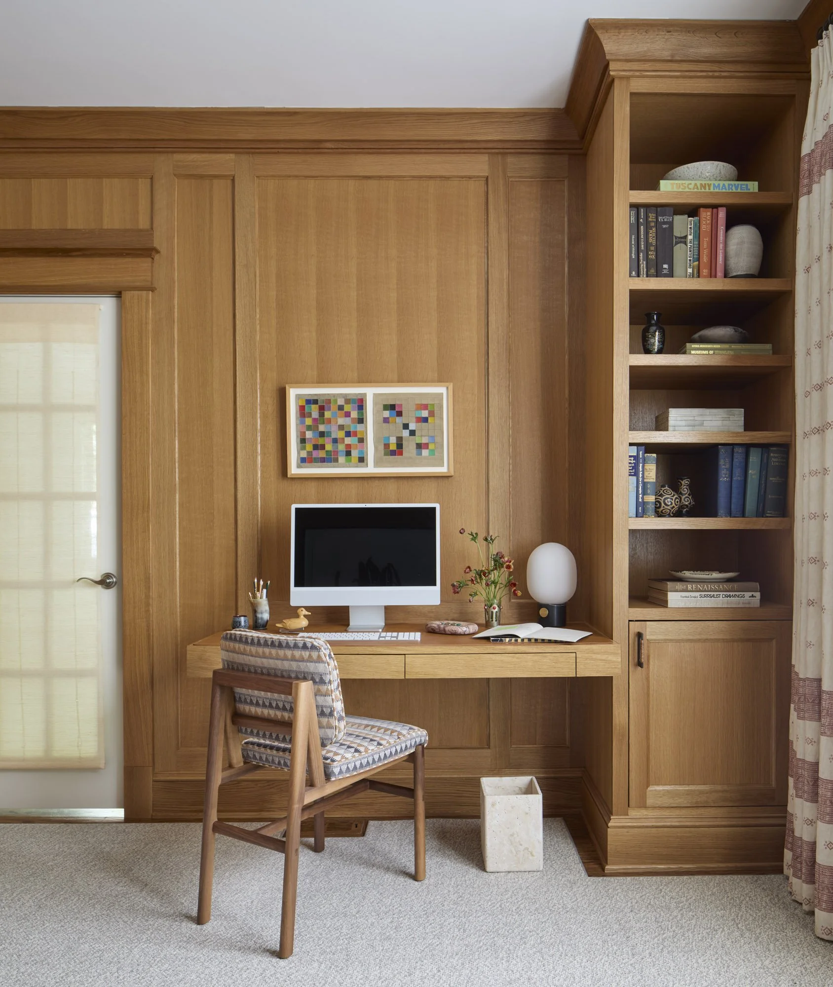 Wood-paneled home office with a wall-mounted desk, patterned chair, abstract artwork, and built-in bookshelves.