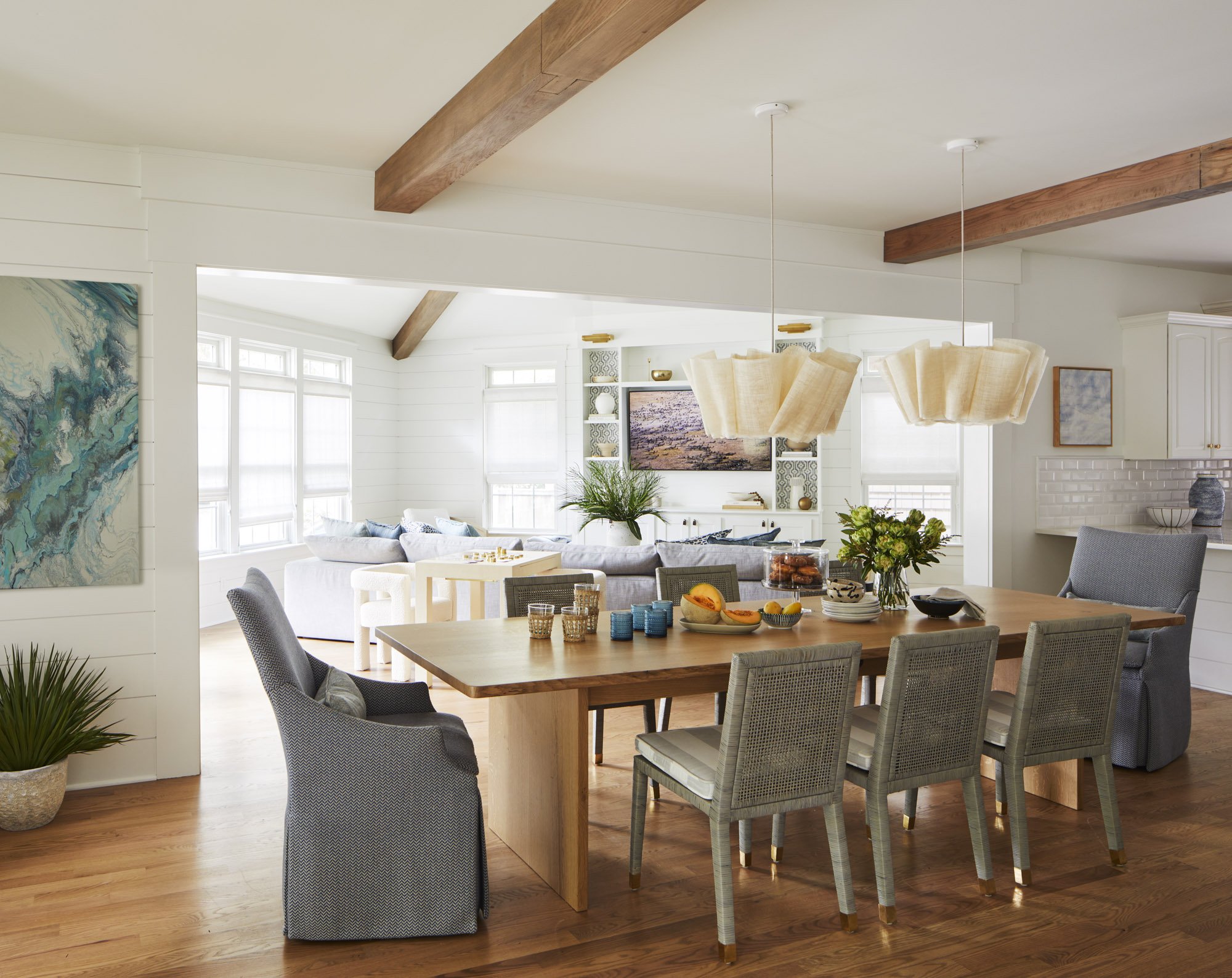 Open dining and living area with exposed wood beams, a large dining table, gray chairs, sculptural pendant lights, and coastal artwork.