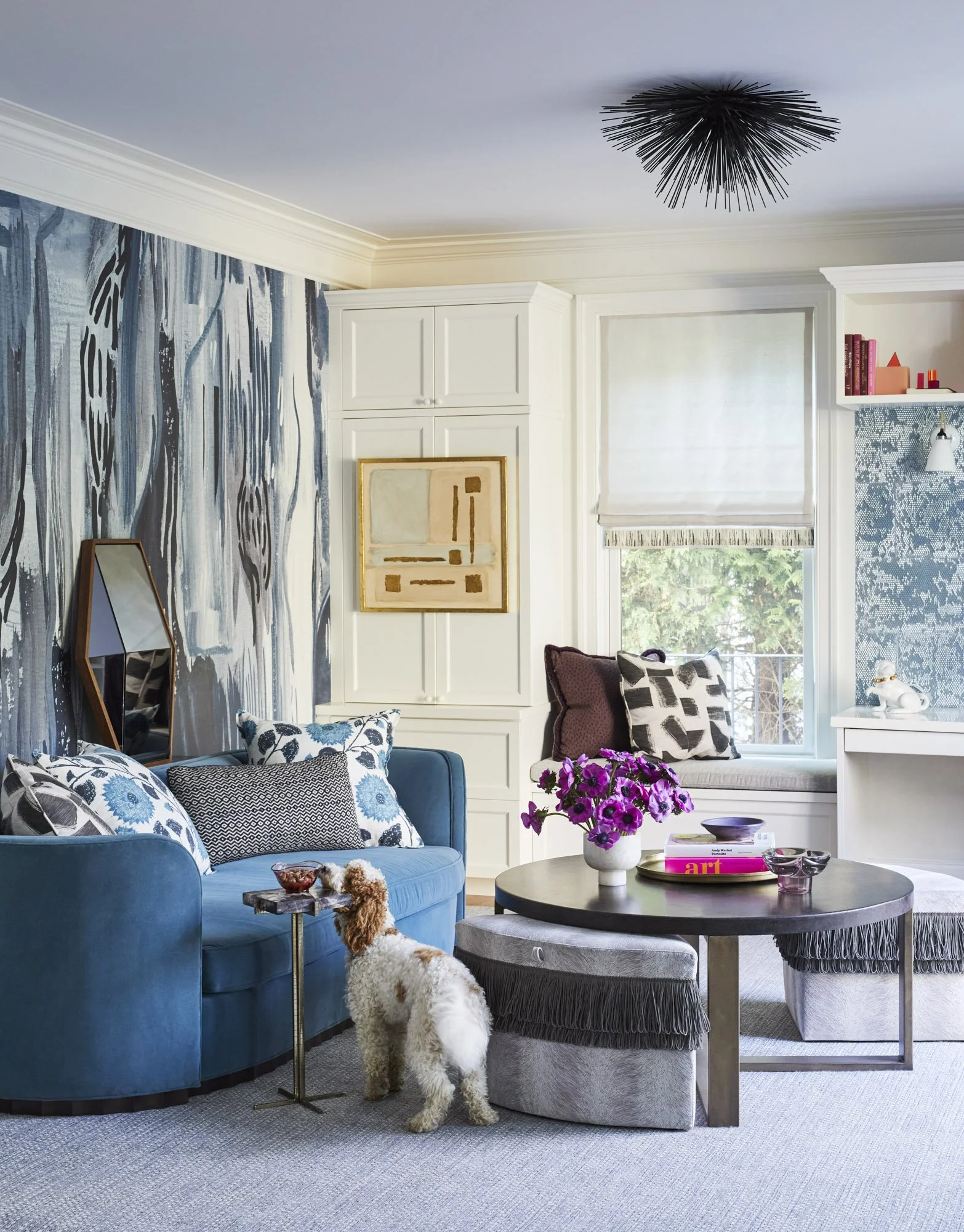 Blue-and-white sitting room with a blue sofa, abstract wallpaper, sculptural chandelier, geometric mirror, and round coffee table.