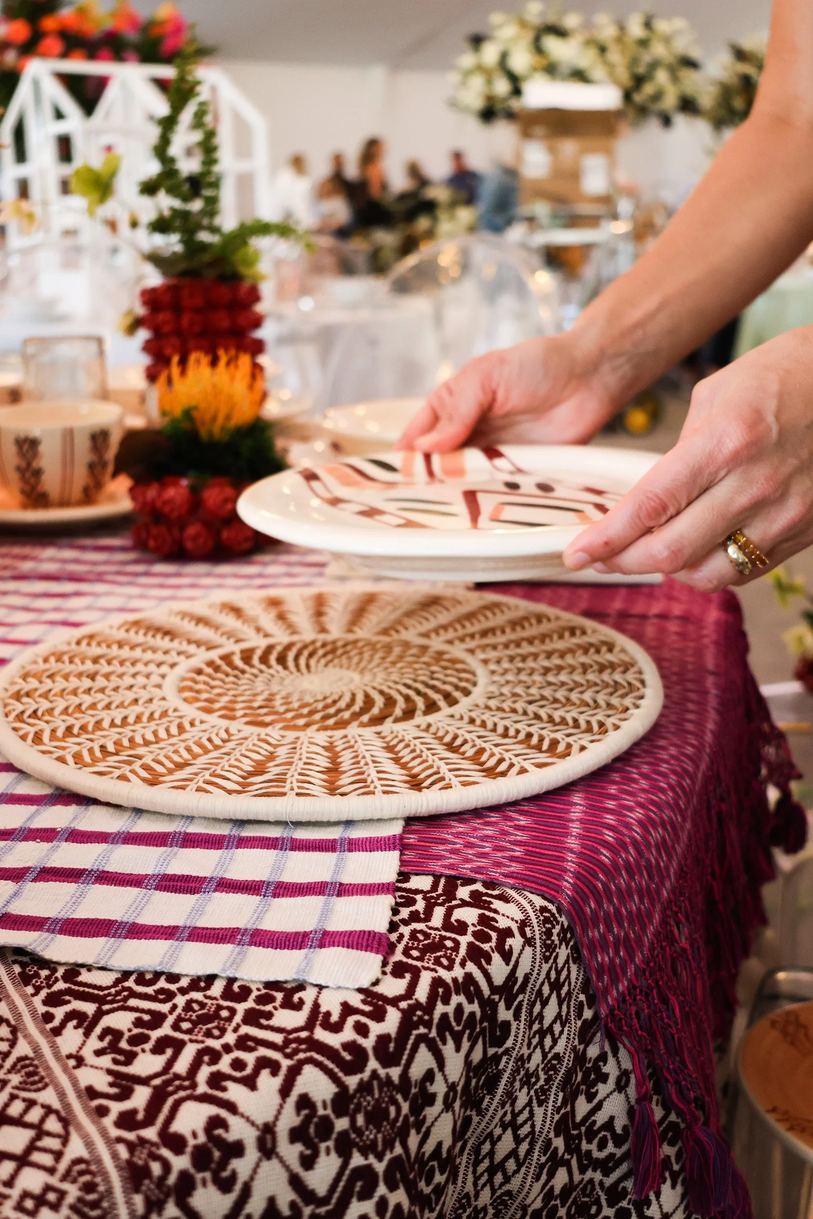 Hands place a hand-painted ceramic plate over a pine needle woven charger and layered RB Curated textiles.