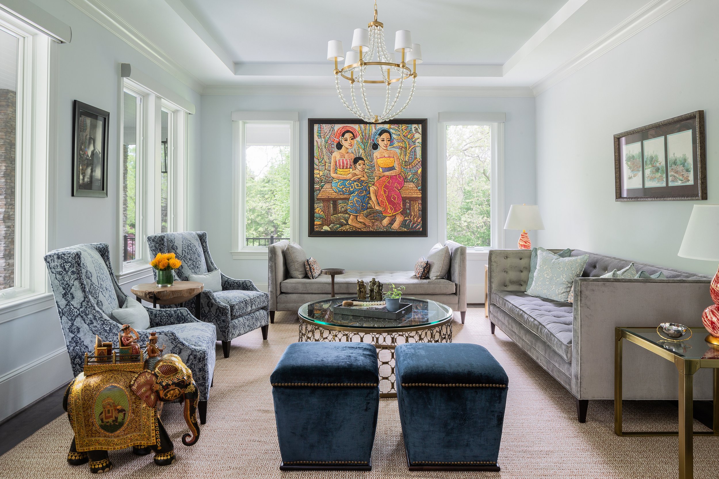 Light blue living room with a large sofa, two armchairs, glass coffee table, elephant sculpture, colorful artwork, and chandelier.