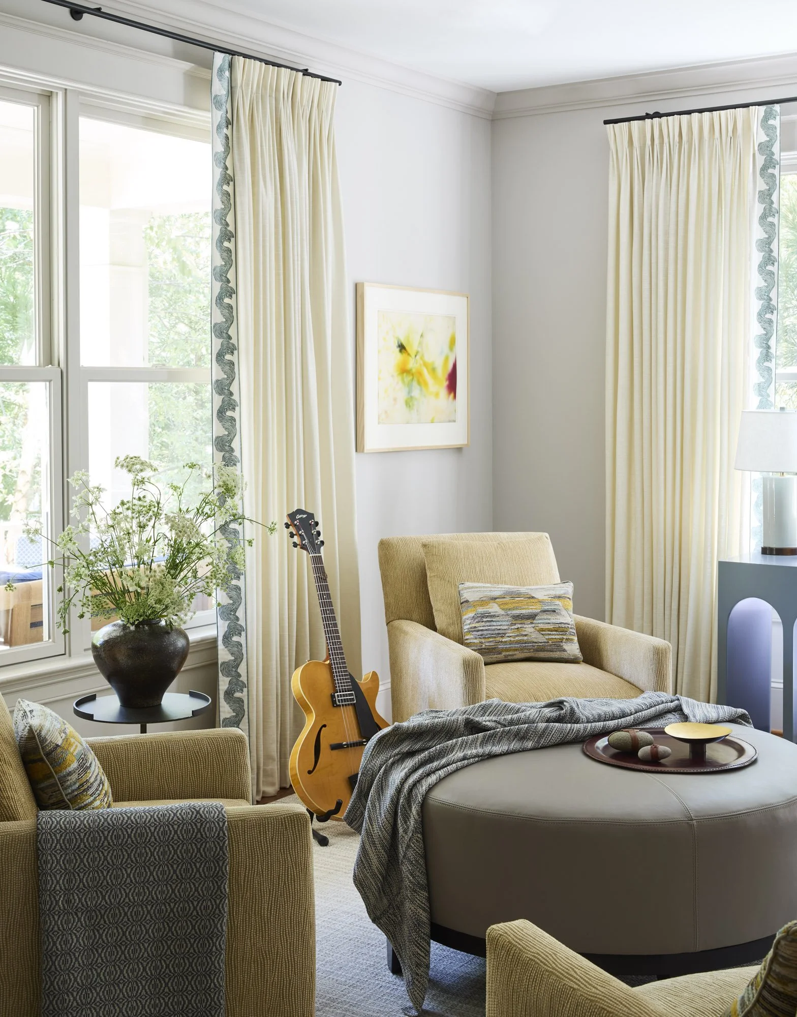 Comfortable music room with beige armchairs, a round ottoman, a wall-mounted guitar, and softly filtered natural light.