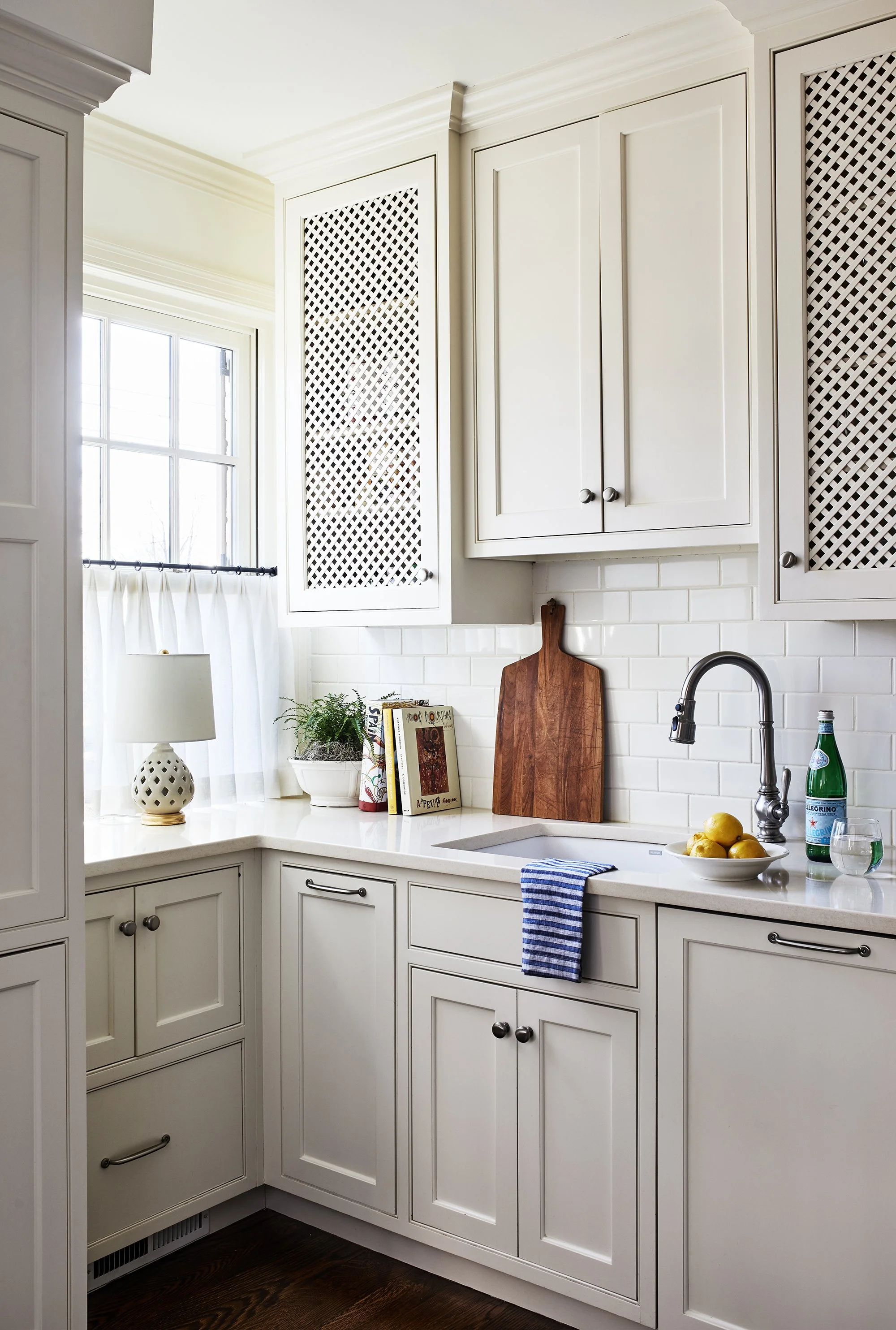 Bright kitchen corner with white lattice-front cabinetry, a sink, polished faucet, cookbooks, lemons, and a window with sheer curtains.