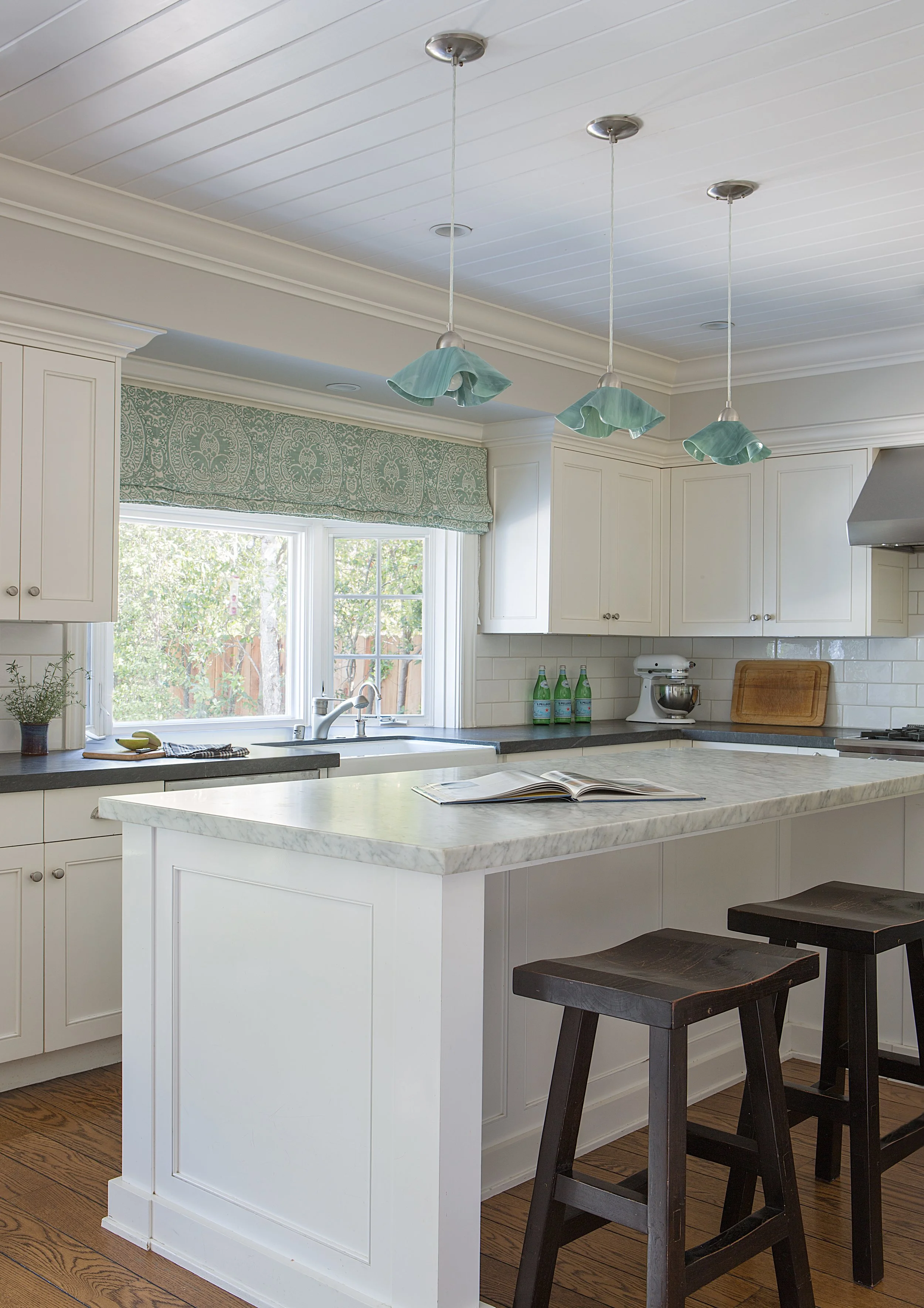 Bright kitchen with white cabinetry, marble countertops, green pendant lights, wood counter stools, and a patterned window shade.