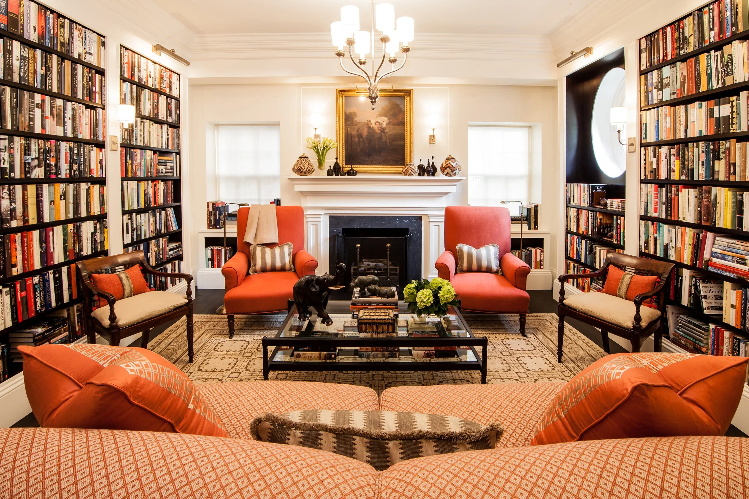 Cozy library living room with book-filled shelves, orange armchairs, a white fireplace, and a glass coffee table.