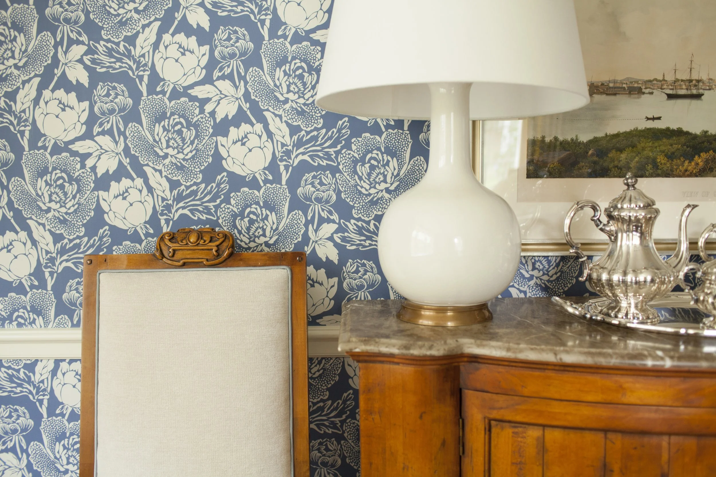 Vintage wood chair and sideboard with a white table lamp and silver tea set against blue floral wallpaper.