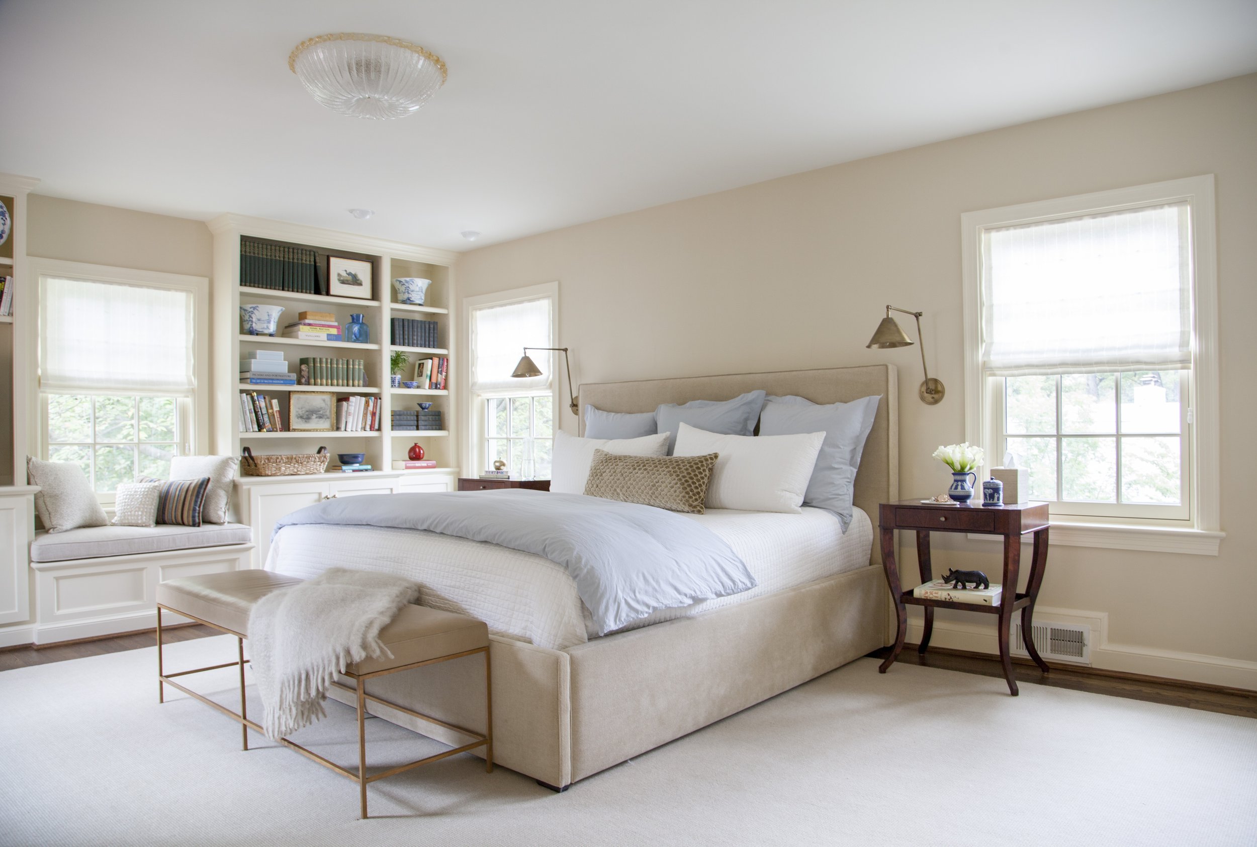 Serene bedroom with a beige upholstered bed, light blue bedding, wall-mounted lamps, built-in bookshelves, and a window seat.