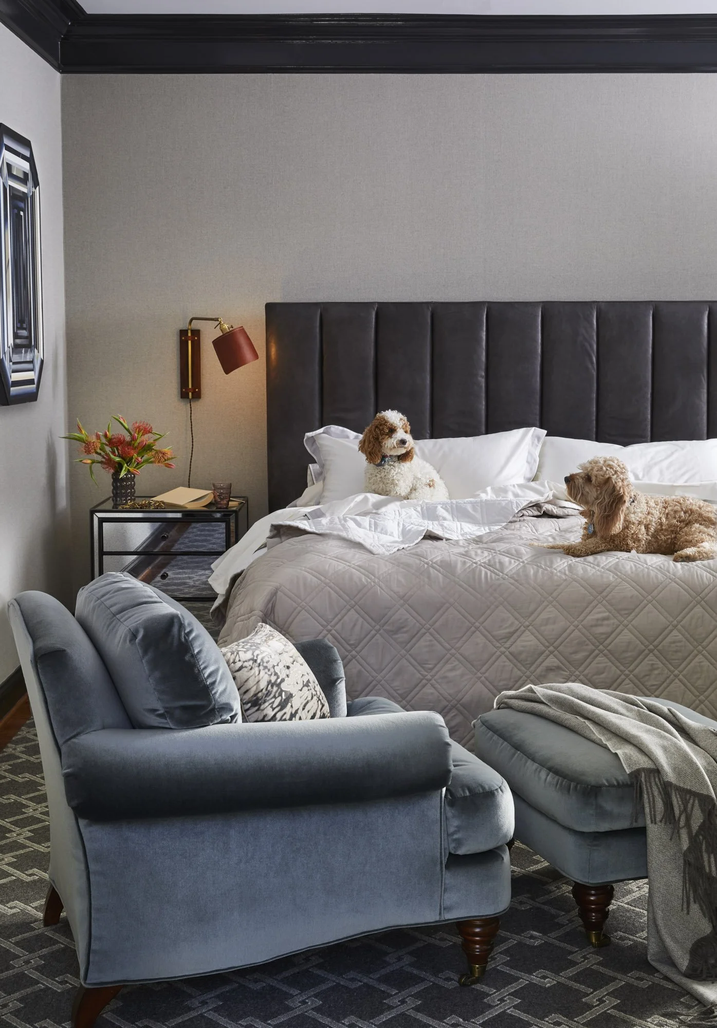 Bedroom with a gray upholstered bed, patterned pillows, blue armchair and ottoman, flowers, and a modern wall light.