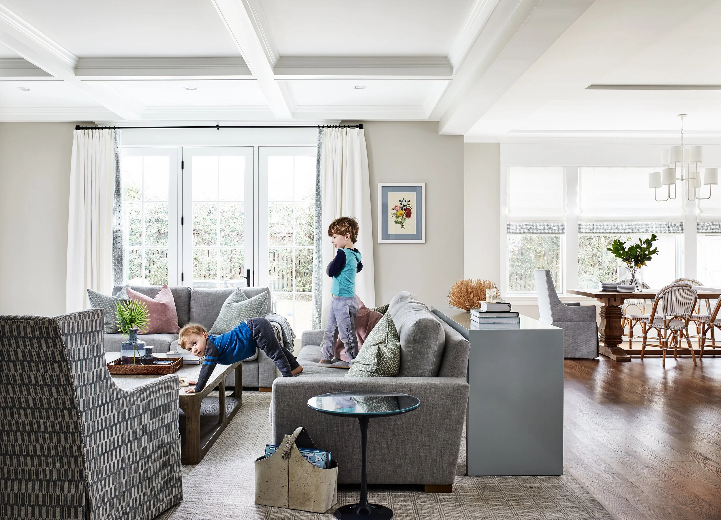 Children playing in a family living room with a gray sofa, armchair, wood coffee table, and dining area in the background.