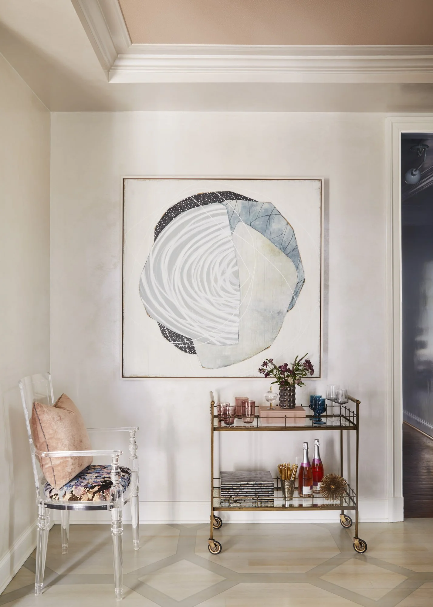 Abstract artwork displayed beside an acrylic chair and a styled bar cart with glassware and flowers.