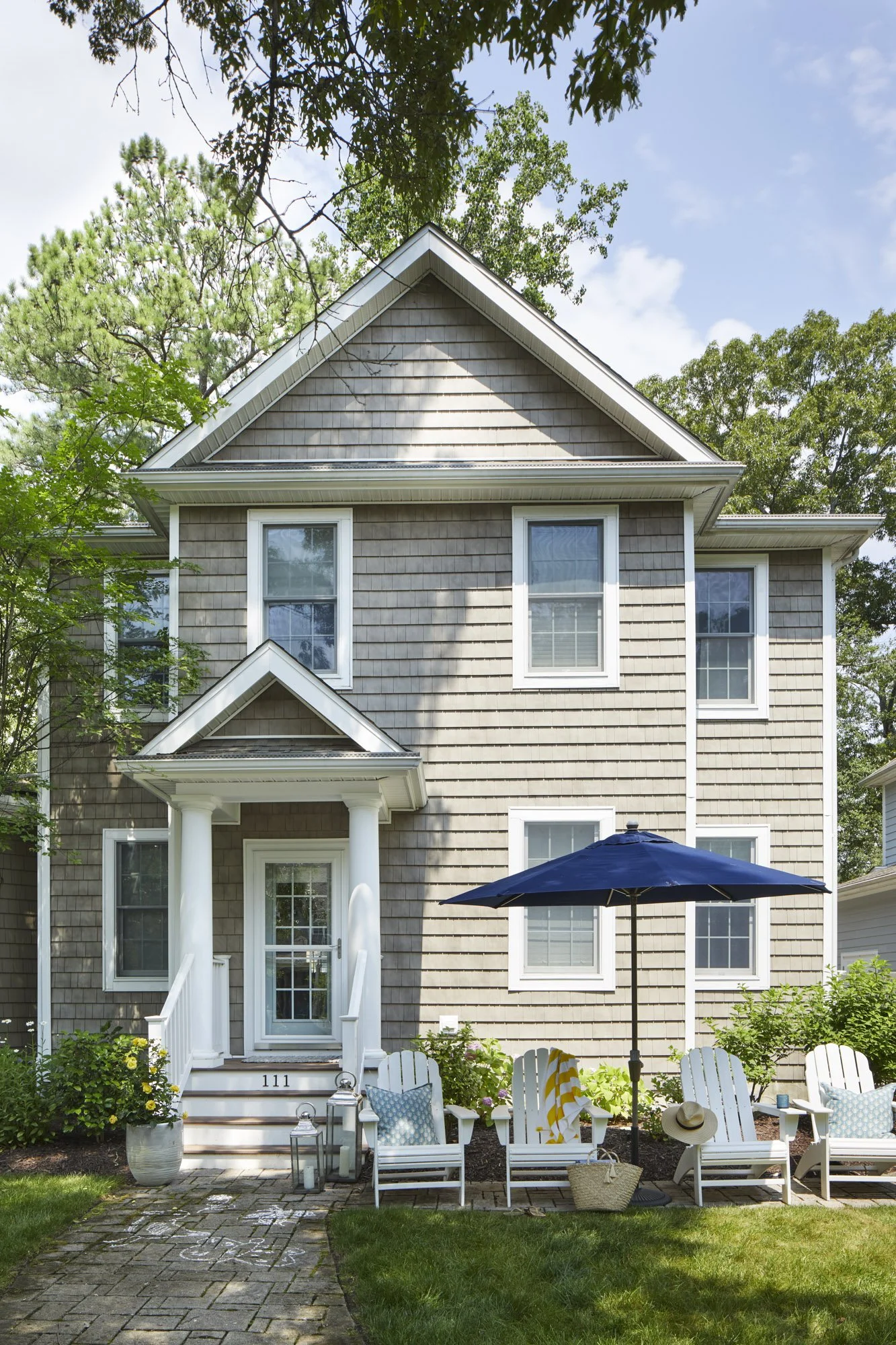 Two-story Rehoboth Beach house with gray shingle siding, a columned porch, Adirondack chairs, and mature trees