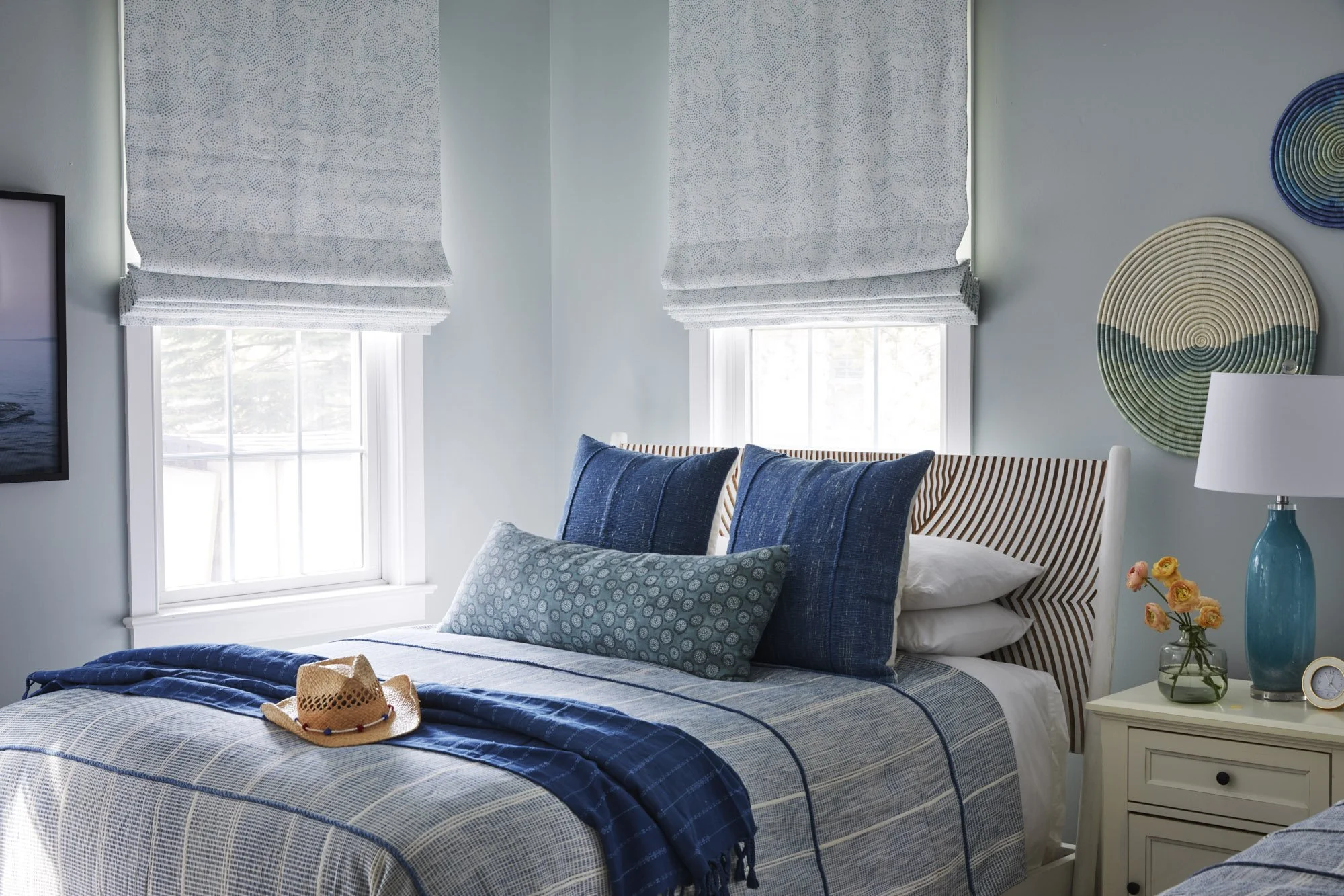Coastal bedroom with blue striped bedding, layered pillows, blue Roman shades, a straw hat, yellow flowers, and circular wall art.