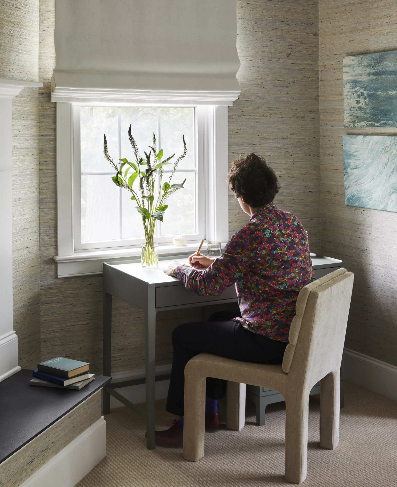 Person writing at a small desk beside a window with textured wallpaper, books, tall branches, and wave artwork.