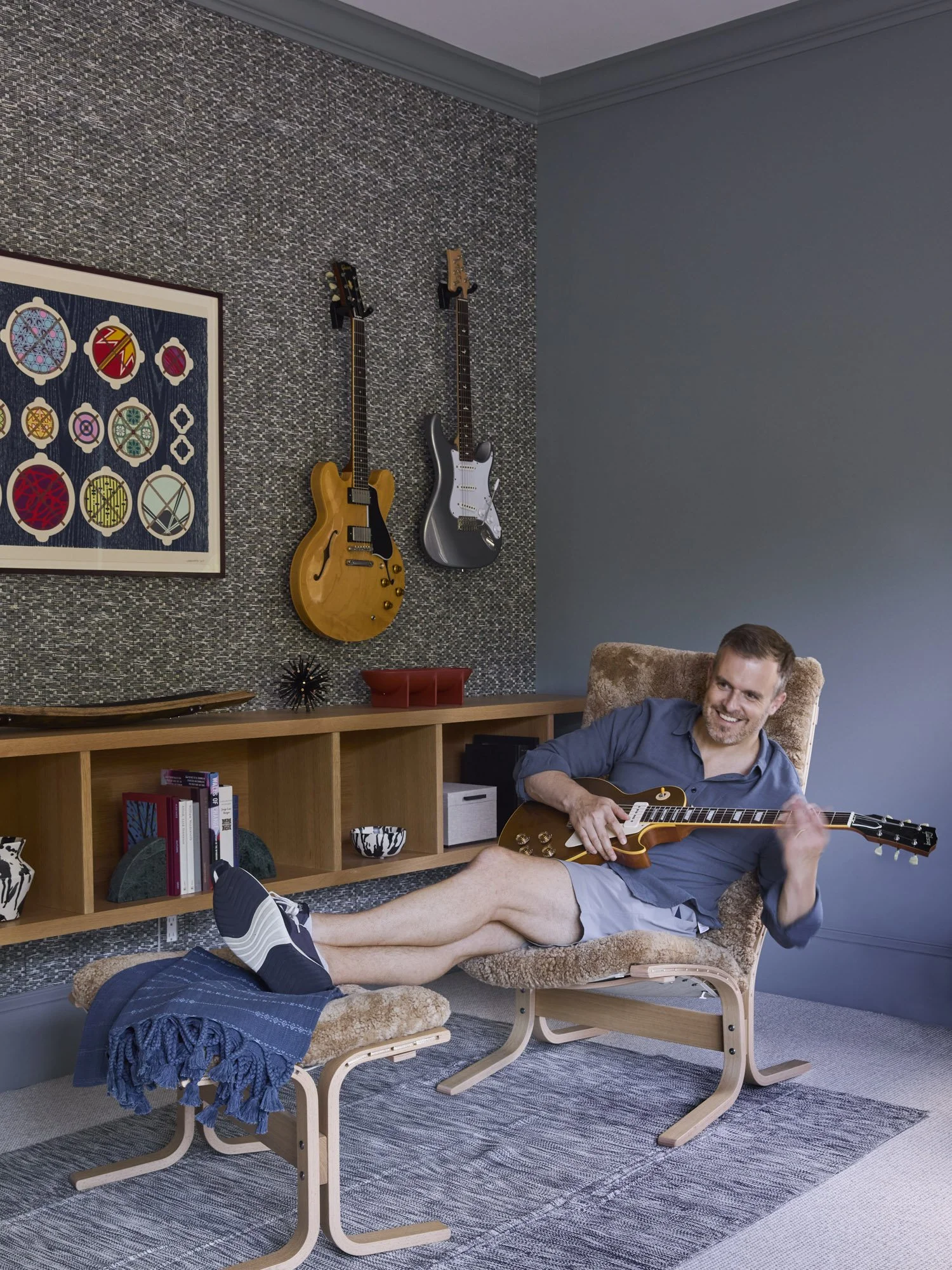 Man plays an electric guitar in a music room with additional guitars and built-in bookshelves.