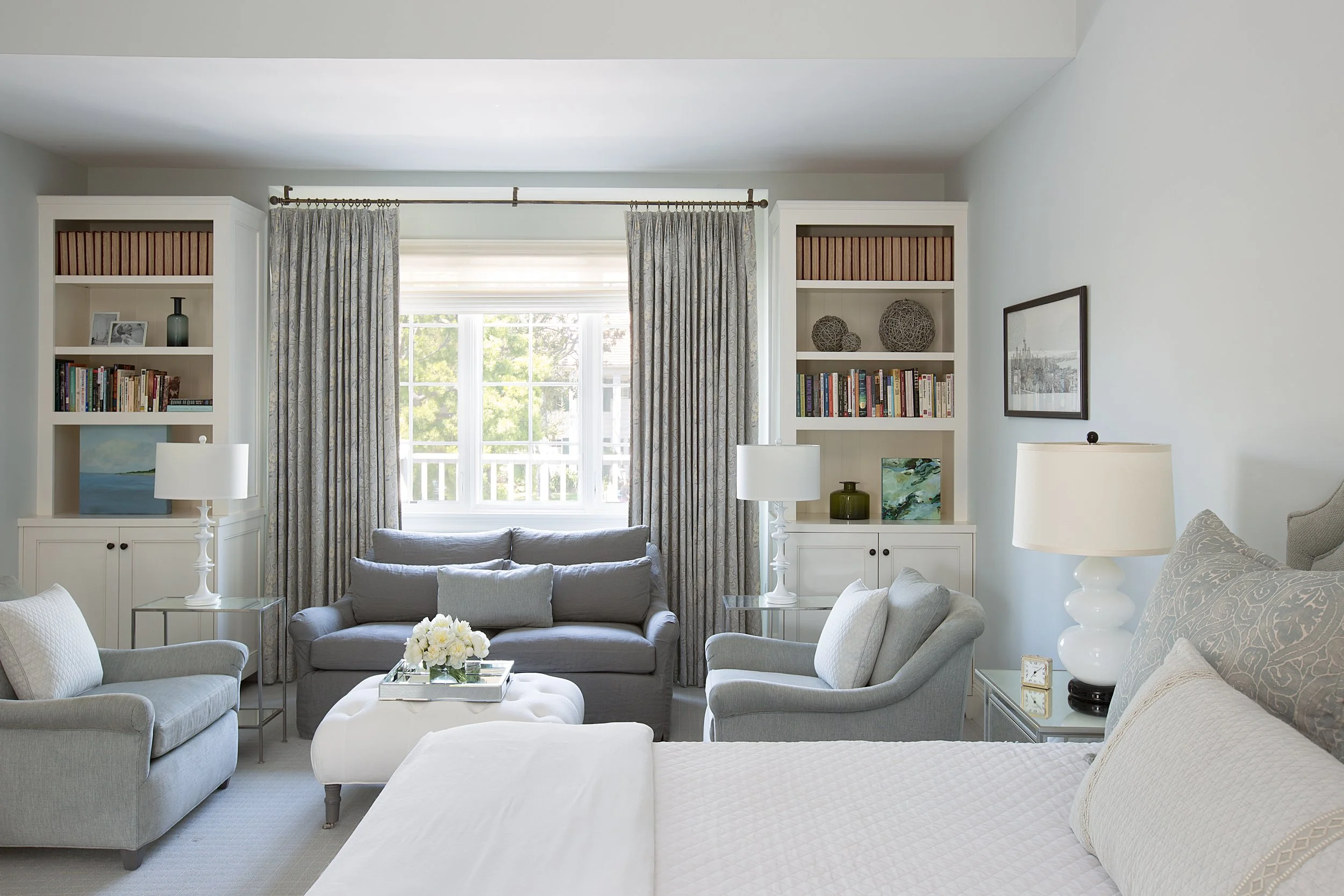 Bedroom with sitting area, a gray sofa, armchairs, white ottoman, built-in bookshelves, large windows, and decorative table lamps.