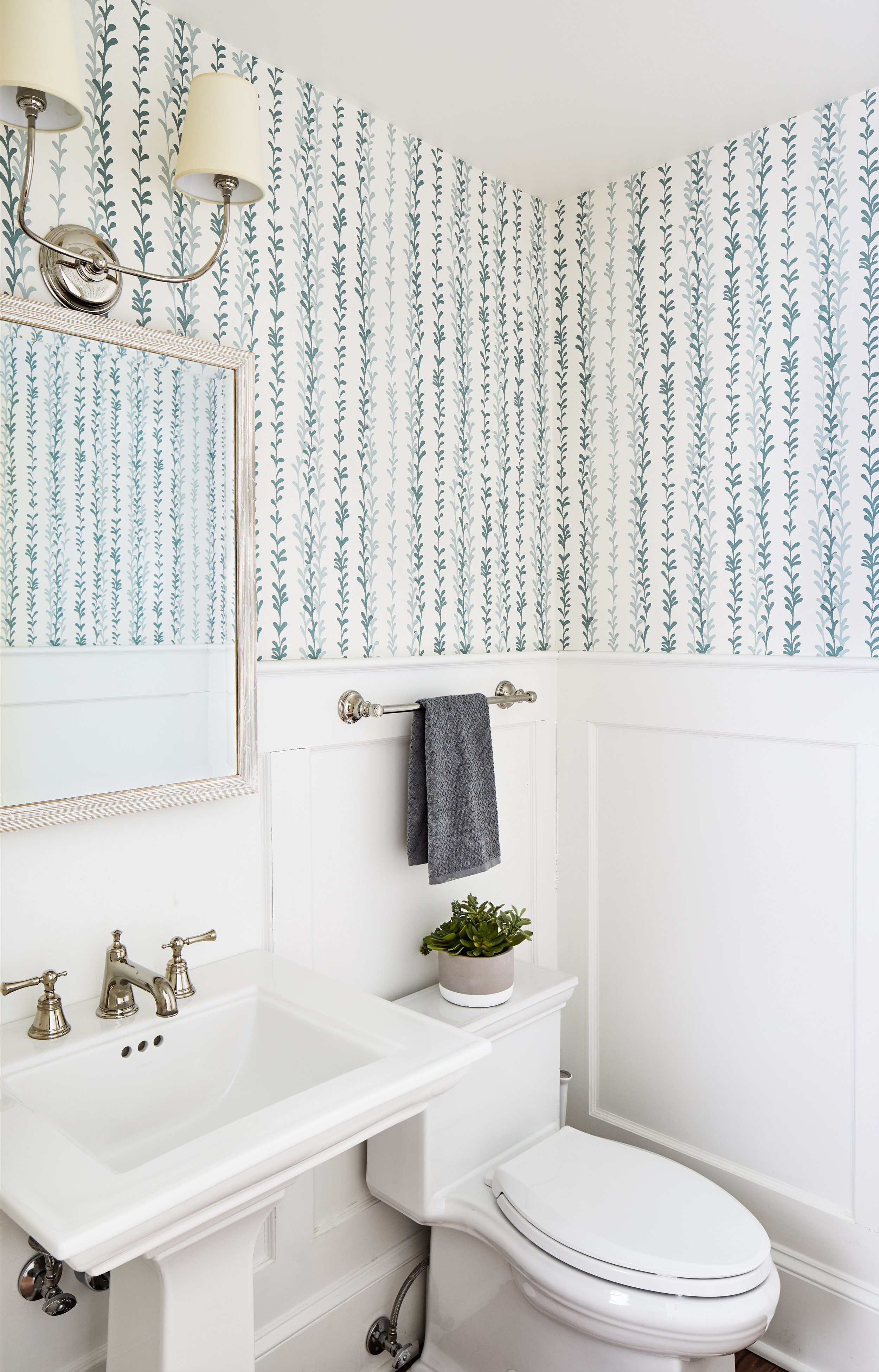 Small powder room with patterned wallpaper, a white pedestal sink, mirror, wall sconces, gray towel, and potted plant.