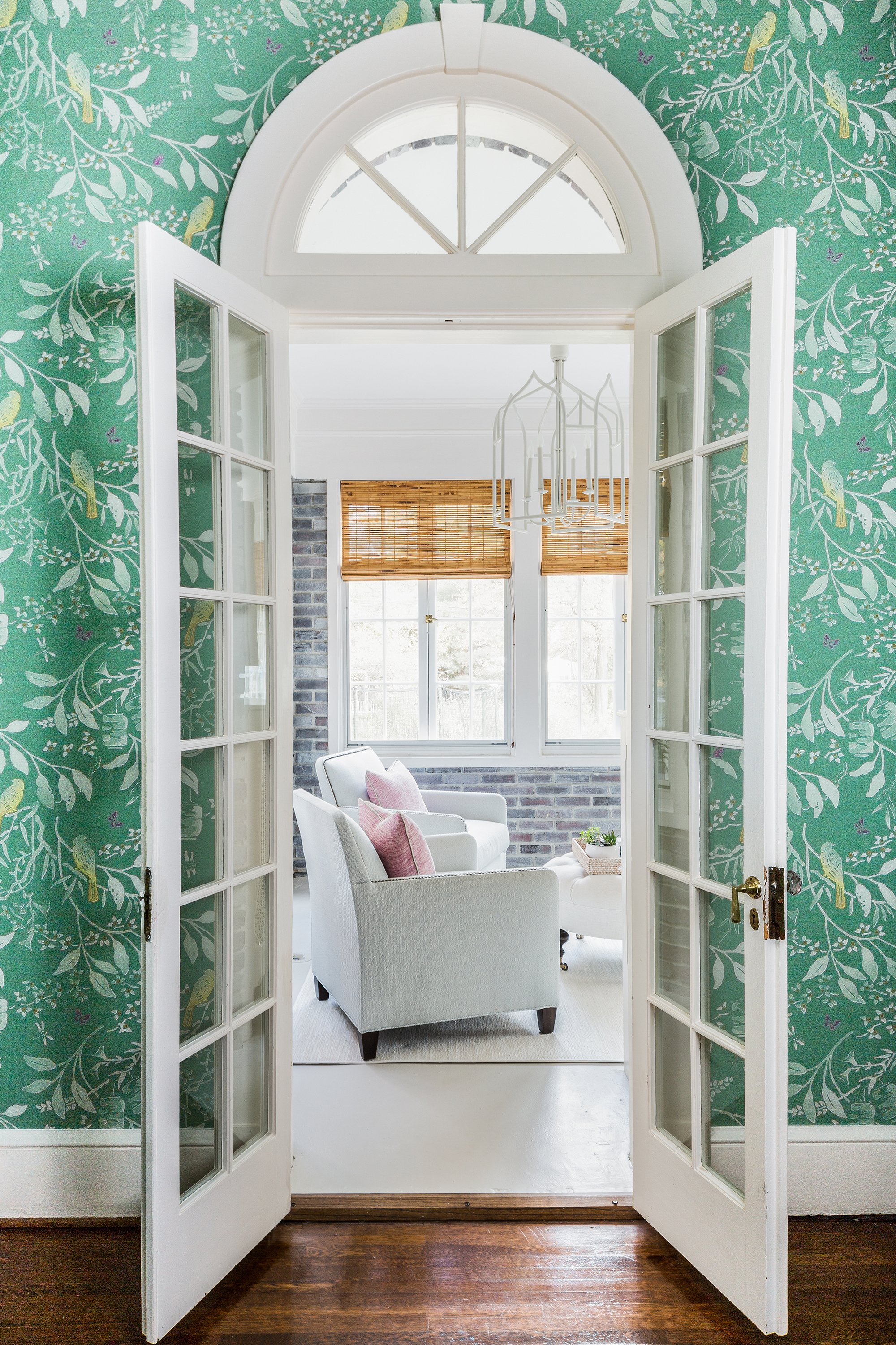 View through open French doors into a sunroom with light blue armchairs, pink pillows, brick walls, bamboo shades, and a white chandelier.