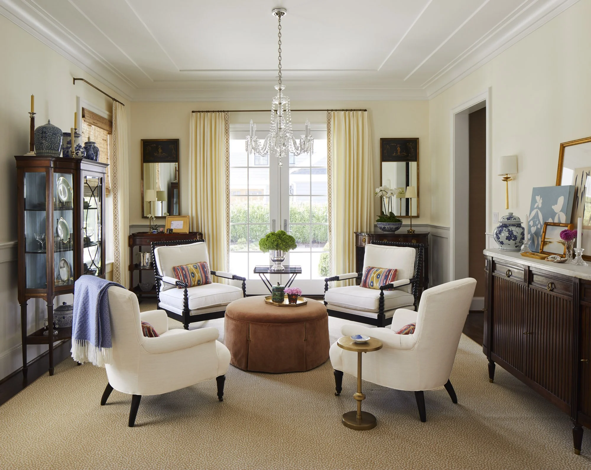 Elegant sitting room with four white armchairs arranged around a round ottoman, mirrored walls, a chandelier, and a glass display cabinet.
