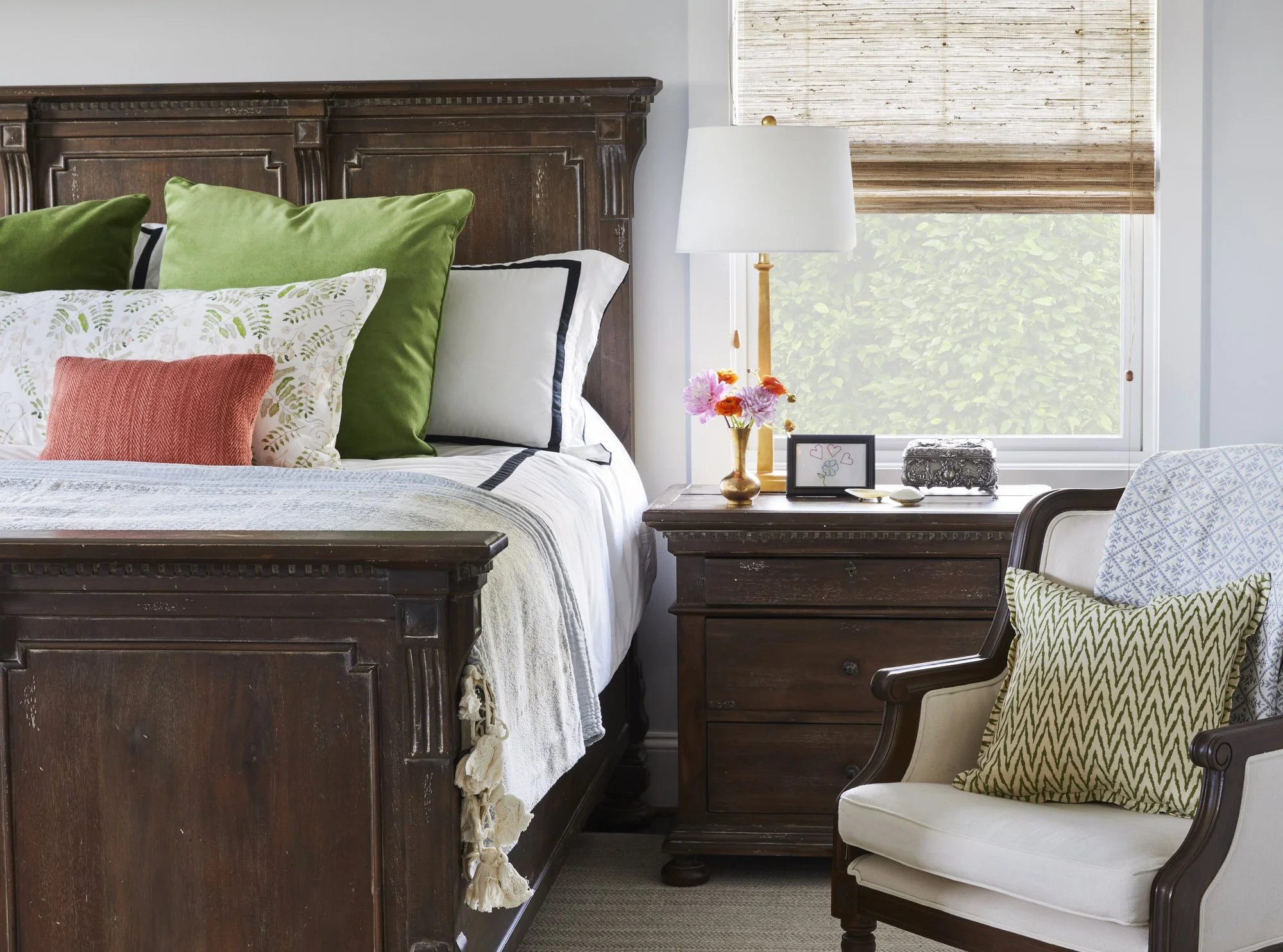 Warm bedroom with a wood bed, green pillows, layered textiles, an upholstered chair, and a bedside table overlooking the greenery.