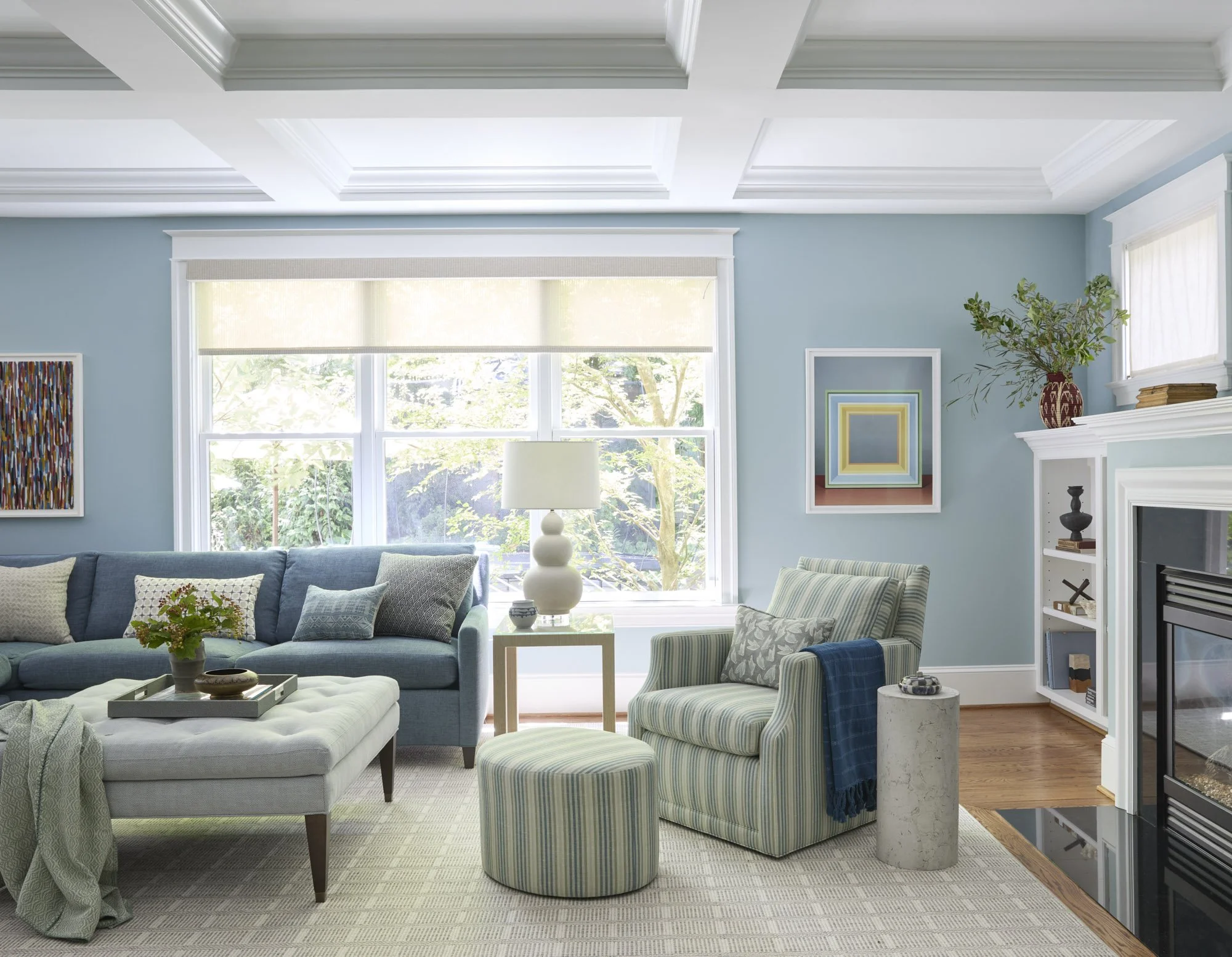 Blue living room with a fireplace, striped armchair, round ottoman, layered artwork, and abundant natural light.