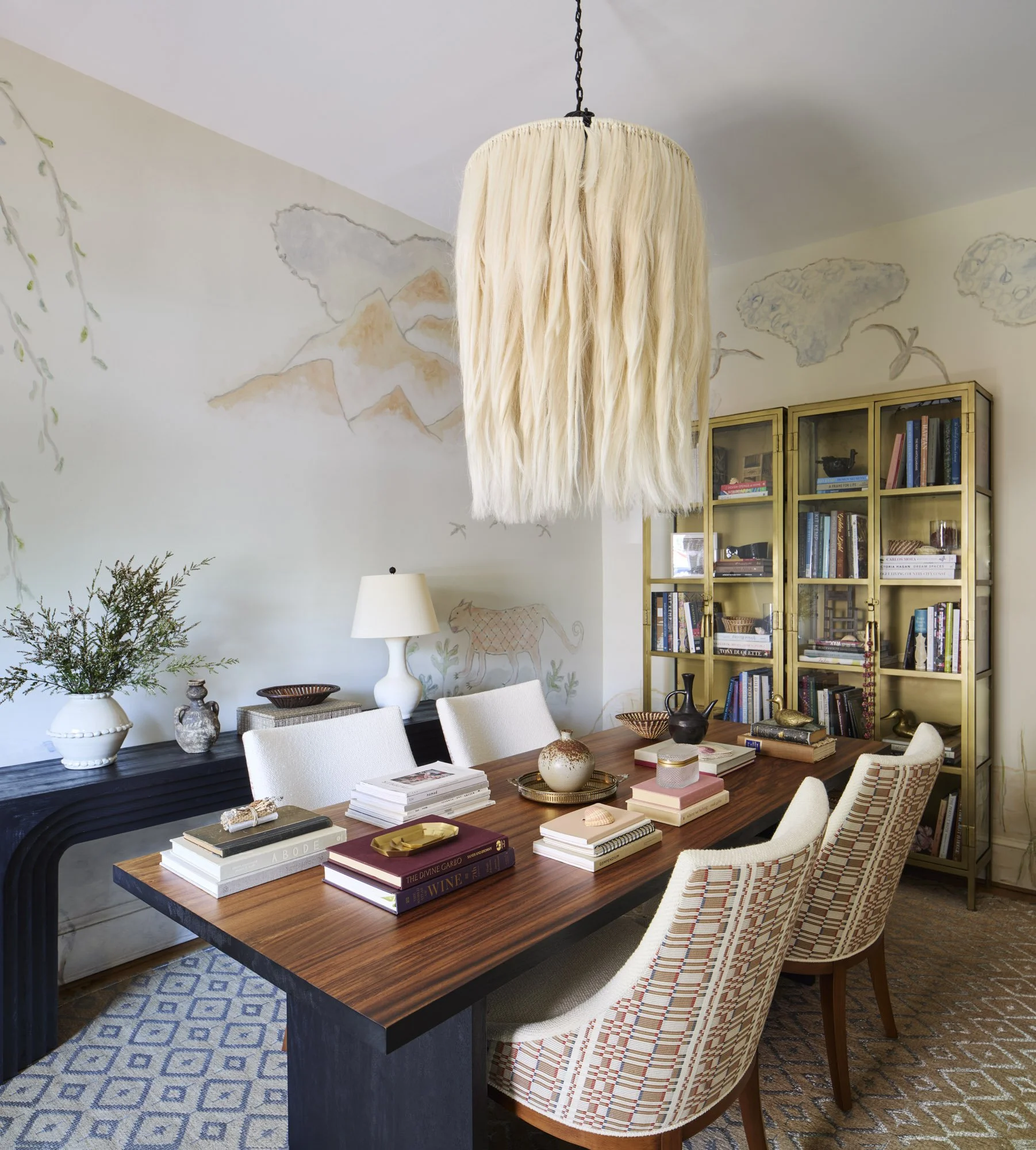 Sculptural feather pendant light above a design studio table with books, collected objects, and patterned chairs.