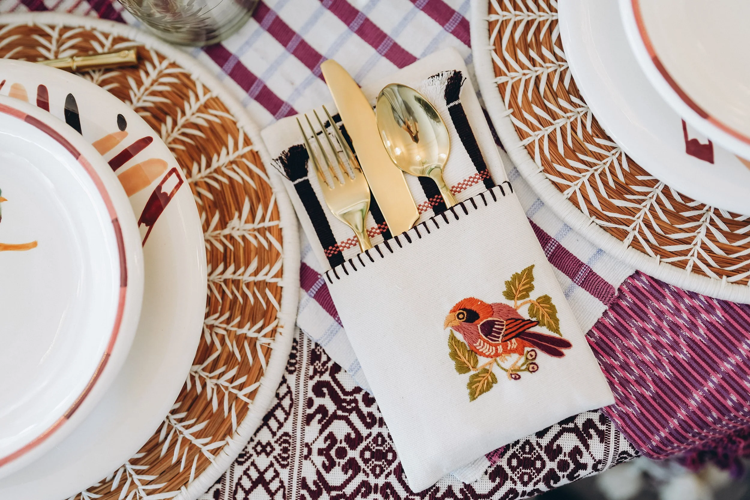 Embroidered bird napkin holding gold flatware beside handmade ceramics, woven chargers, and layered textiles.