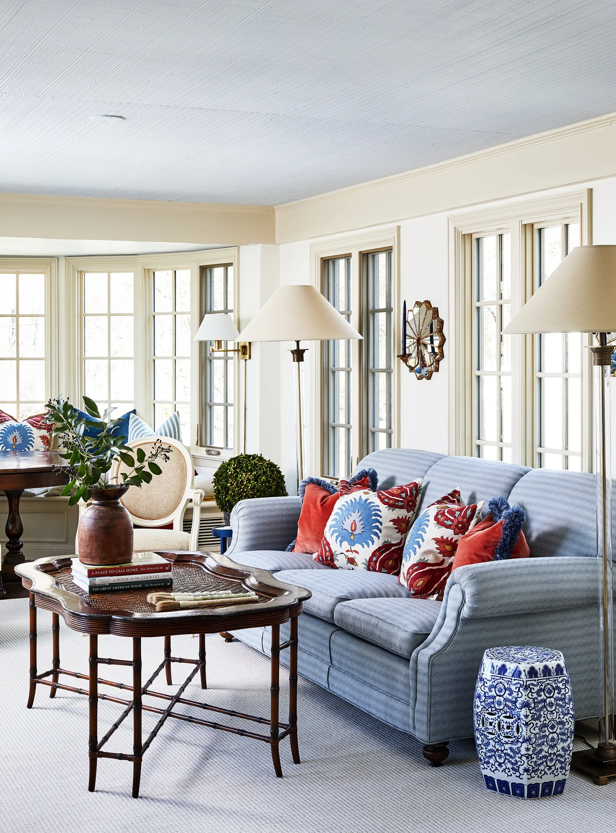 Traditional living room with a blue striped sofa, colorful pillows, wood coffee table, ceramic garden stool, and large windows.