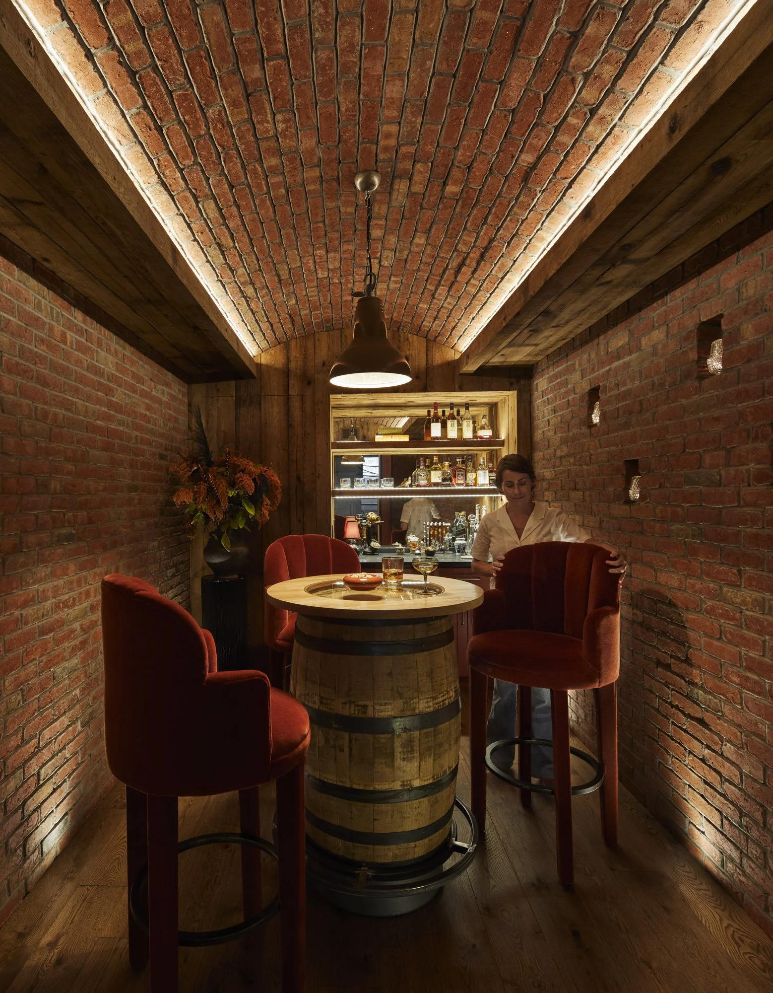 Hidden 1920s-inspired speakeasy with brick walls, a barrel table, red velvet chairs, pendant lighting, and a custom bar.
