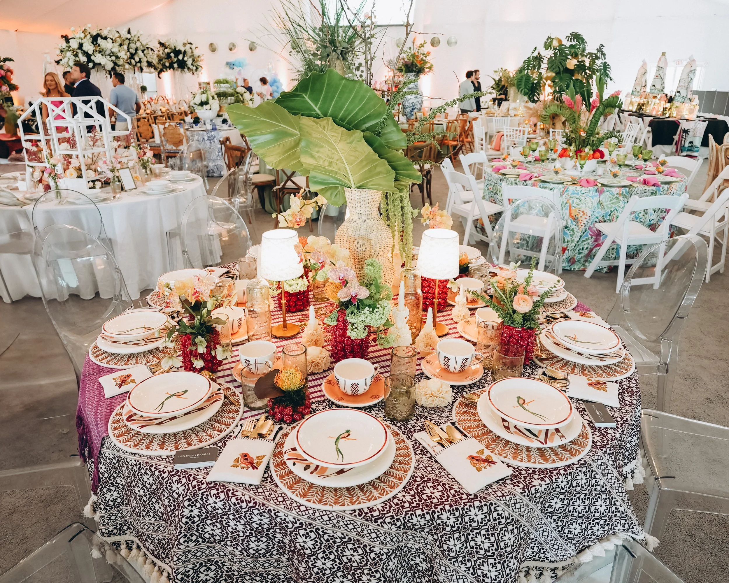 Naples Tables event display featuring handmade ceramics, woven chargers, patterned textiles, table lamps, and a dramatic leafy centerpiece.