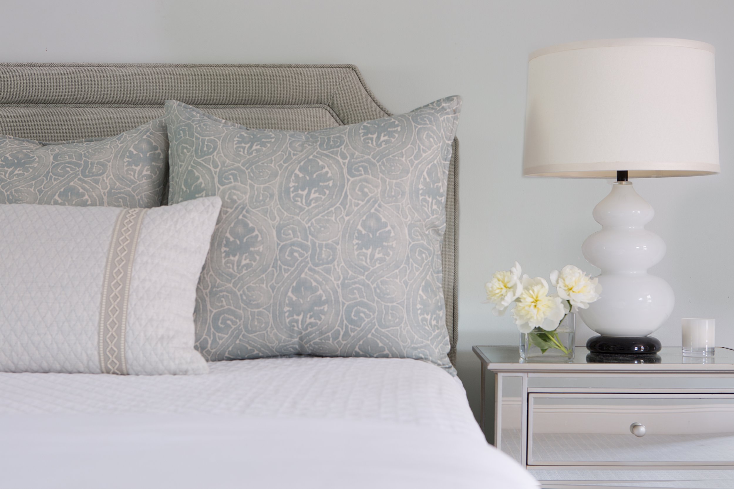 Bedroom detail with layered patterned pillows, a mirrored nightstand, white table lamp, and fresh flowers.