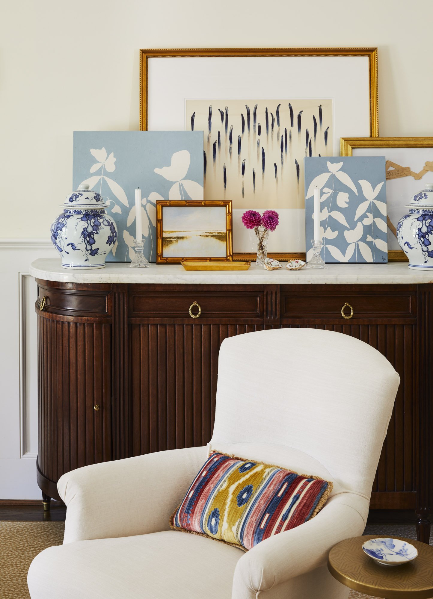 White armchair with a colorful patterned pillow beside a wood sideboard styled with blue-and-white ceramics, artwork, and pink flowers.