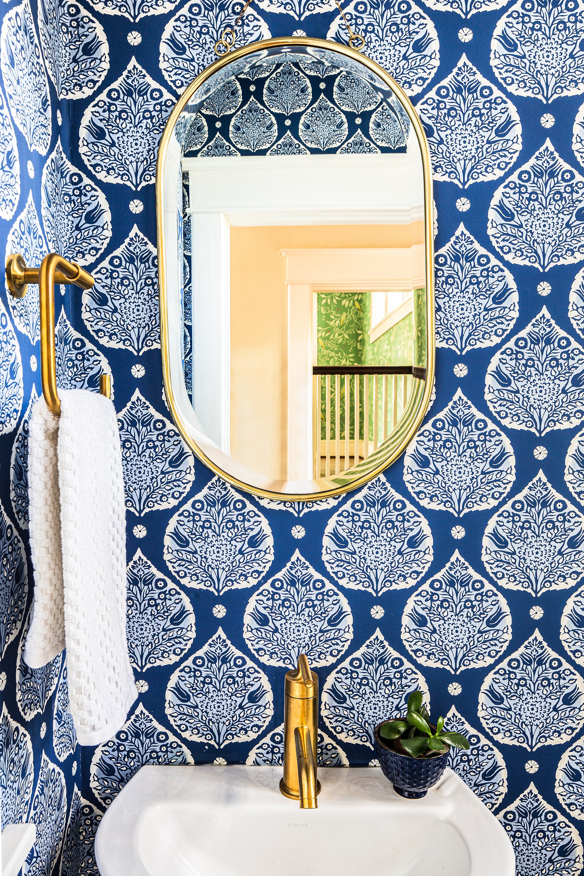 Powder room with blue-and-white floral wallpaper, a gold-framed oval mirror, brass faucet, and white hand towel.