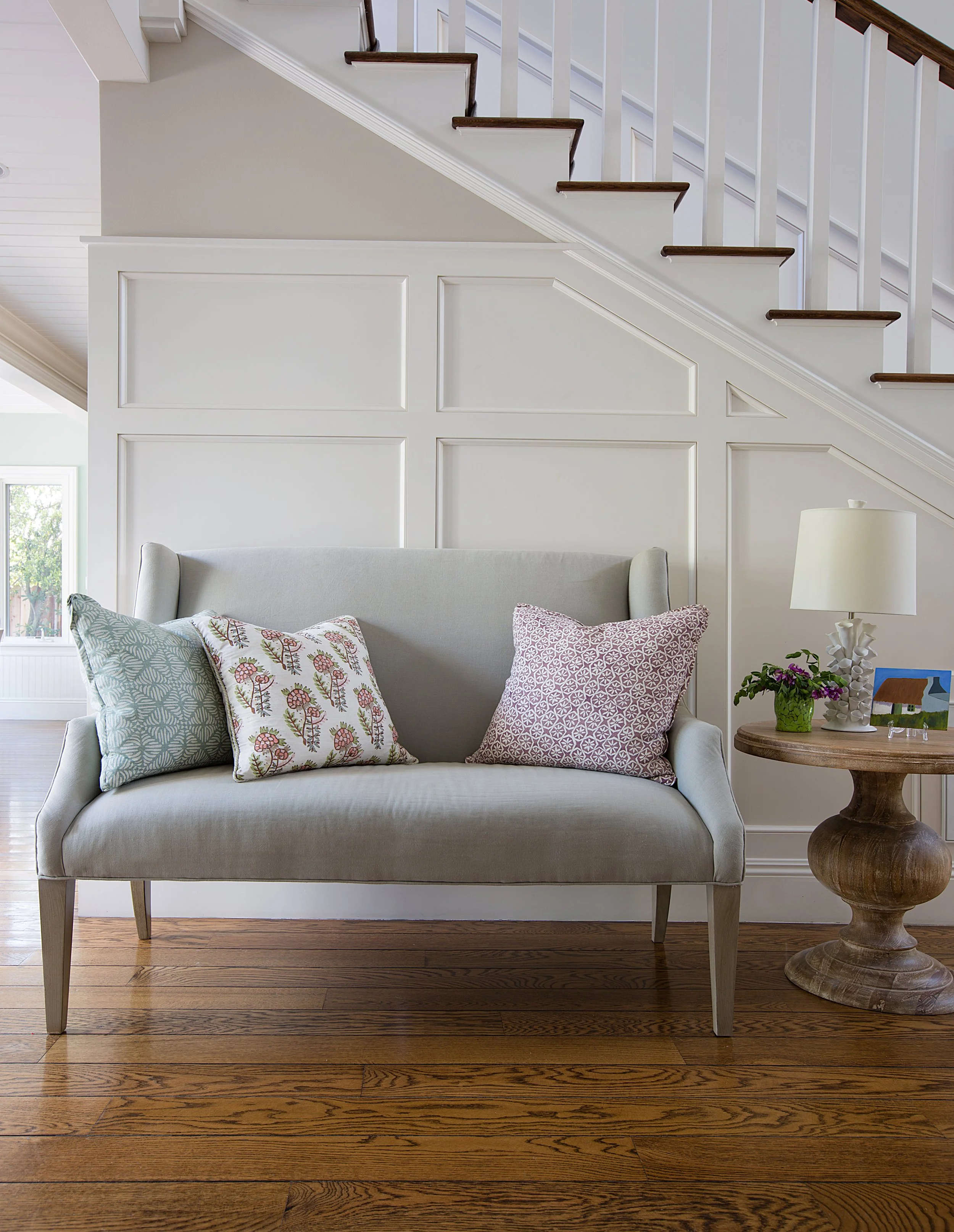 Light gray loveseat with patterned pillows beneath a staircase, beside a round wood table, lamp, and potted plant.